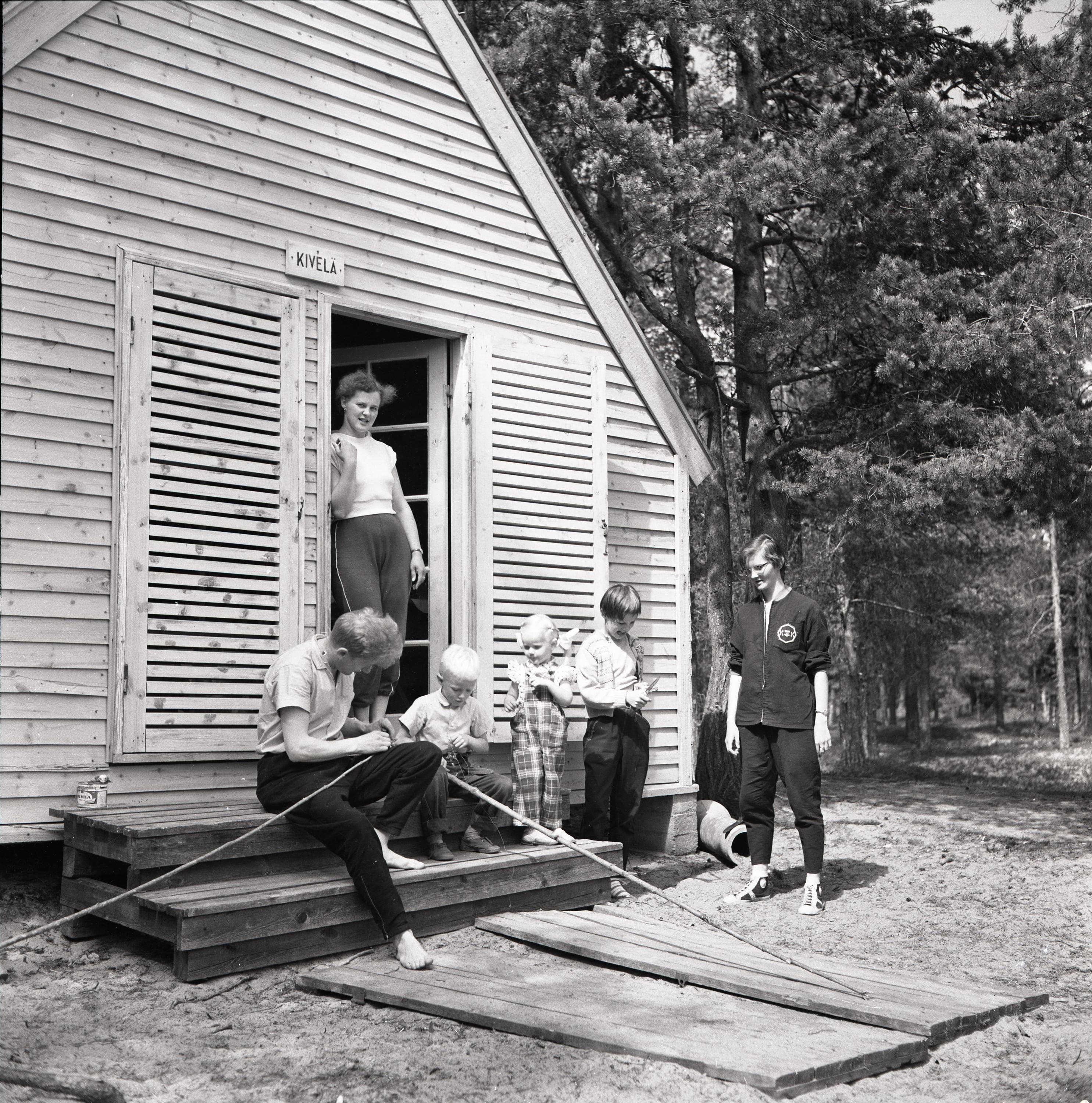 Image: Okänd / Kainuu museo On the left side, a small holiday cottage with people in front of it. On the right side, a forest.