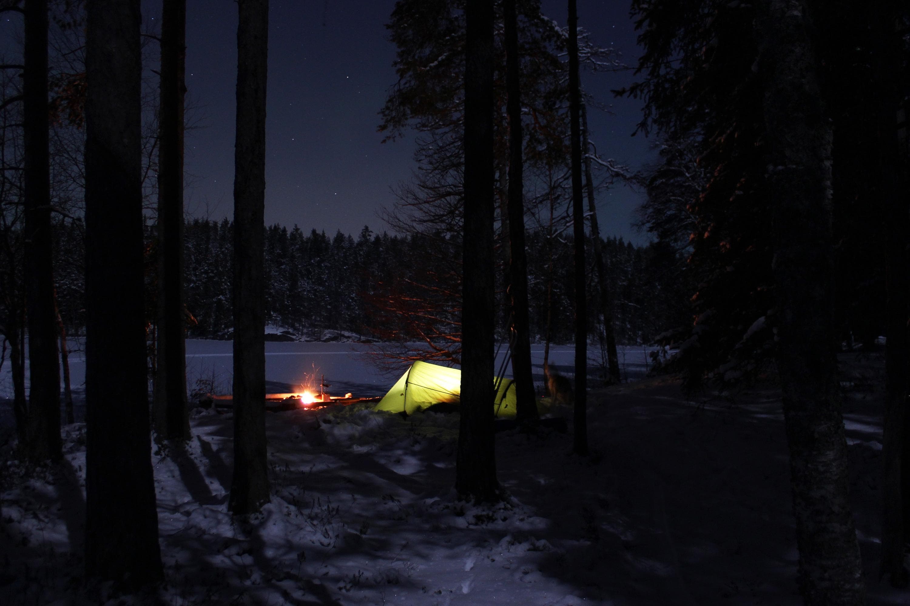 Image: Minna Jakosuo By the lakeshore, there is a tent and a campfire. It is winter and dark. Light comes from the campfire and the glow from inside the tent.