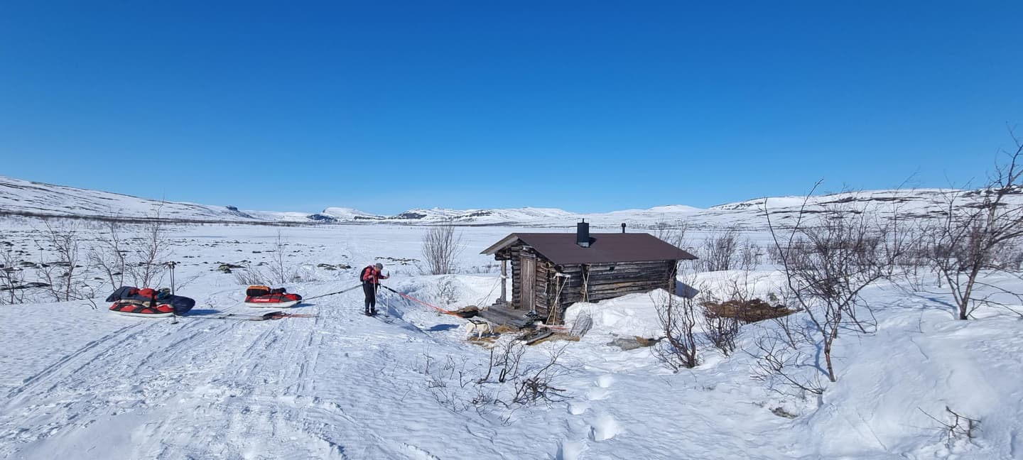 Bild: Annu Tuohiluoto En skidåkare med pulka och hund vid en liten timmerstuga. Två andra pulkor är i närheten. Himlen ovanför fjällen är blå och molnfri.