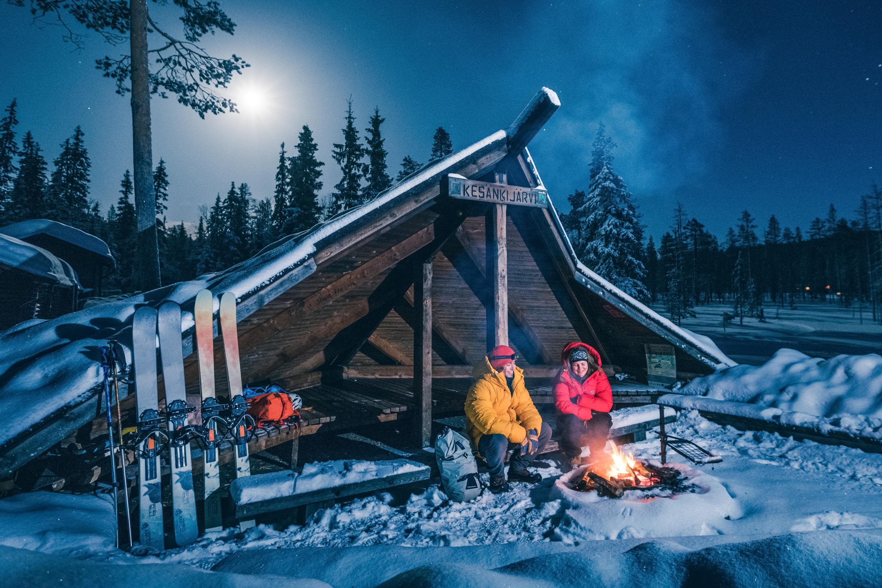 Image: Rami Valonen Two hikers sit at a lean-to shelter in the evening twilight on a winter night. A campfire burns in front of the shelter. The moonlight illuminates the scene.