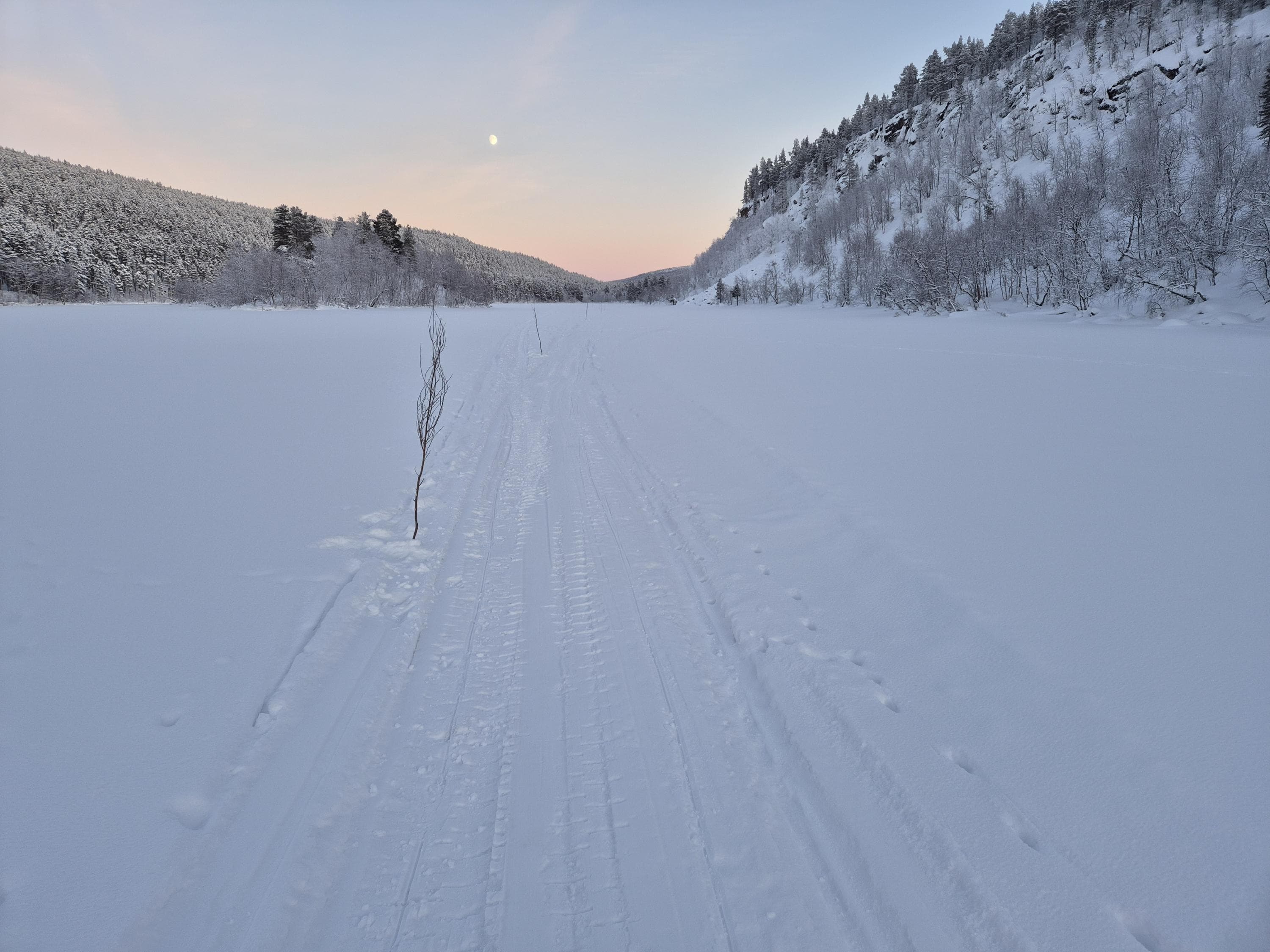 Kuva: Alex Kopf Erämainen joki on jäässä ja ympäröivä metsä lumen peitossa. Jokijäätä halkoo risukepein merkitty moottorikelkan ura.