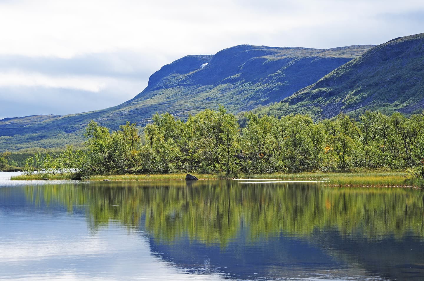Image: Seija Olkkonen Behind the lake, a birch forest can be seen, with a tall mountain looming in the background.