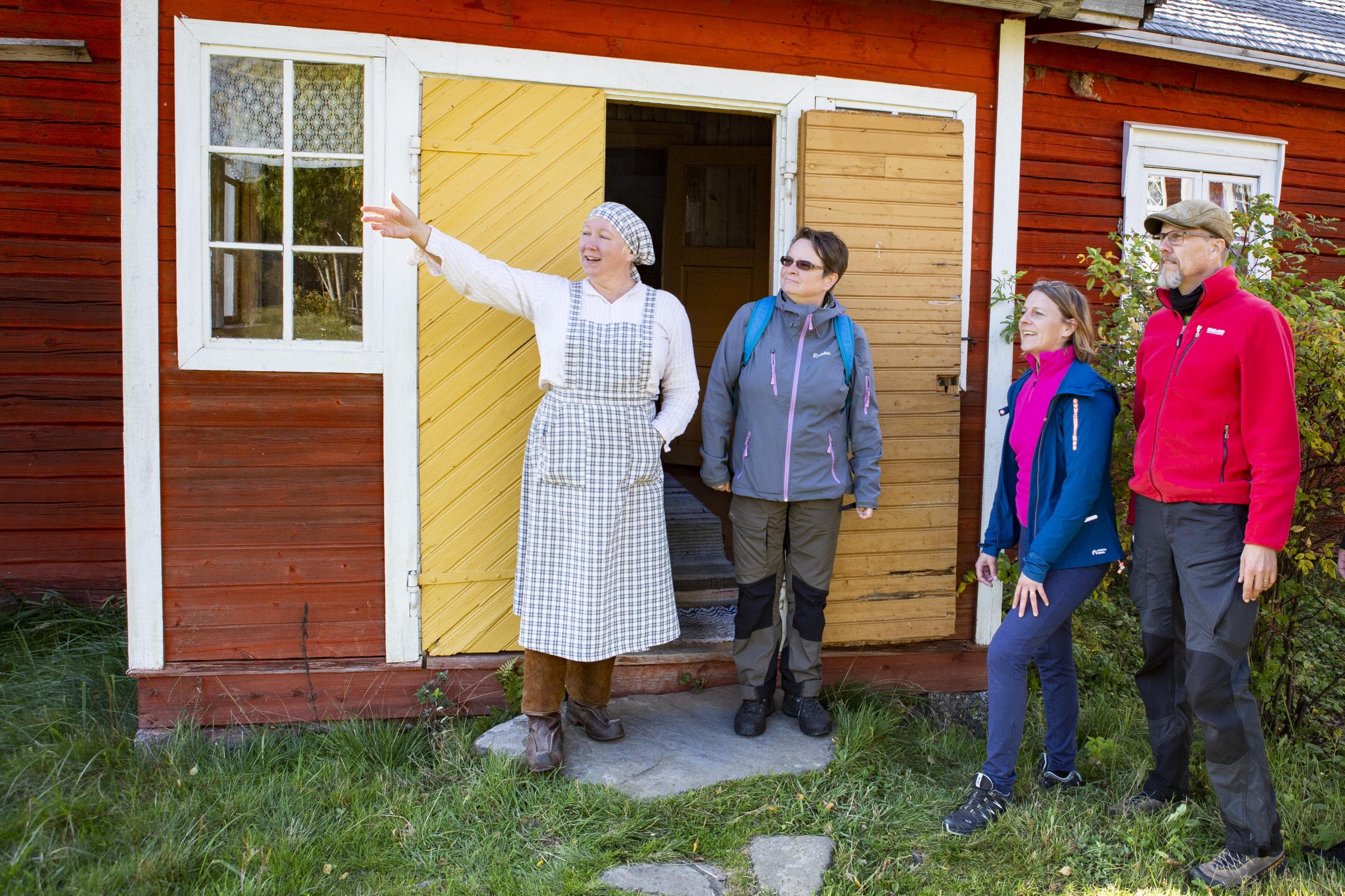Kuva: Salla Penttilä Vanhan ajan vaatteisiin pukeutunut henkilö esittelee henkilöille pihaa punaisen rakennuksen edessä.