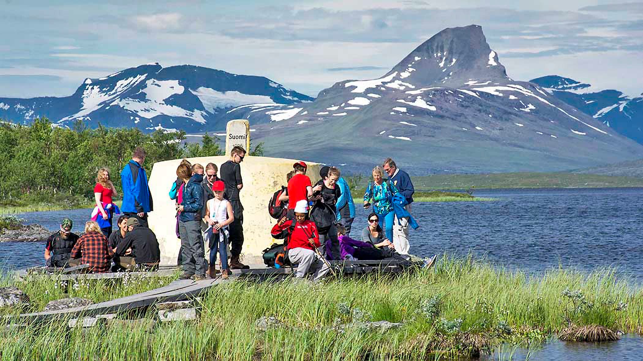 Image: Seija Olkkonen People around a low, round concrete structure. On top of the structure, a sign reads "Suomi." In the background, there is a lake, a birch forest, and fells.