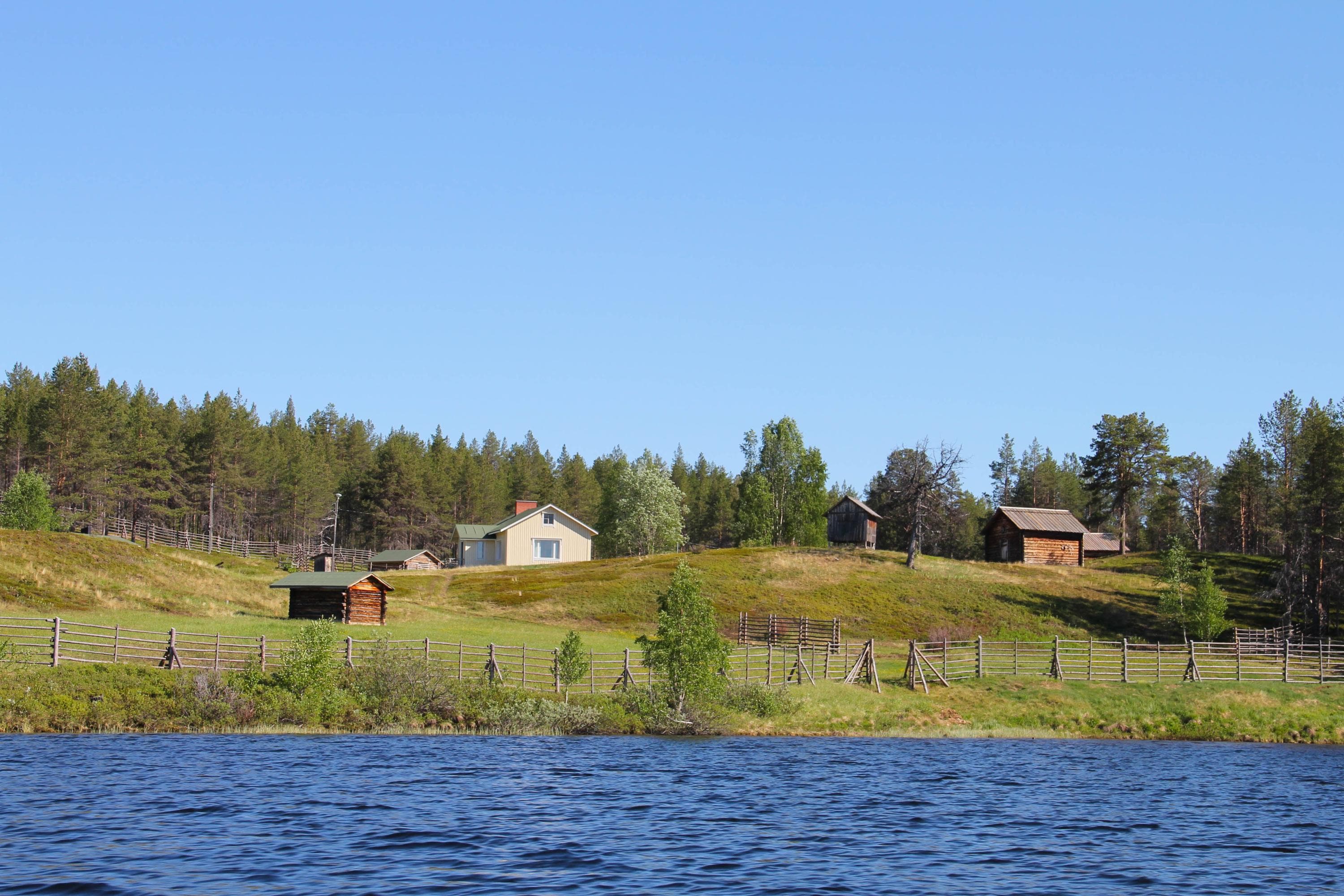 Kuva: Kirsi Ukkonen Asuinkenttä kuvattuna joelta. Niityn takana on asuintalo ja piharakennuksia.Taivas on pilvetön.