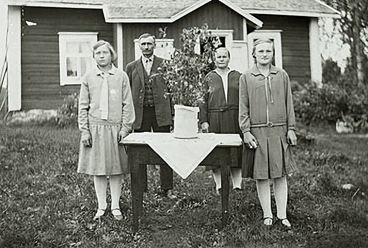 Four people are standing in front of the cottage around a table in an old photograph.
