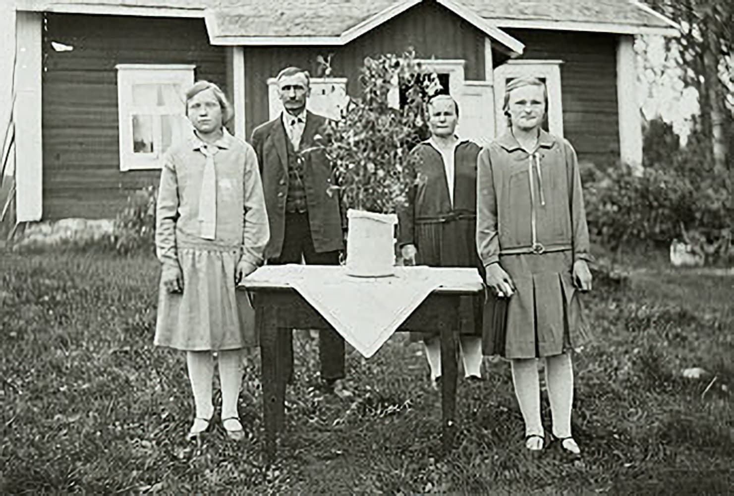 Fyra personer står framför en stuga runt ett bord på ett gammalt fotografi.