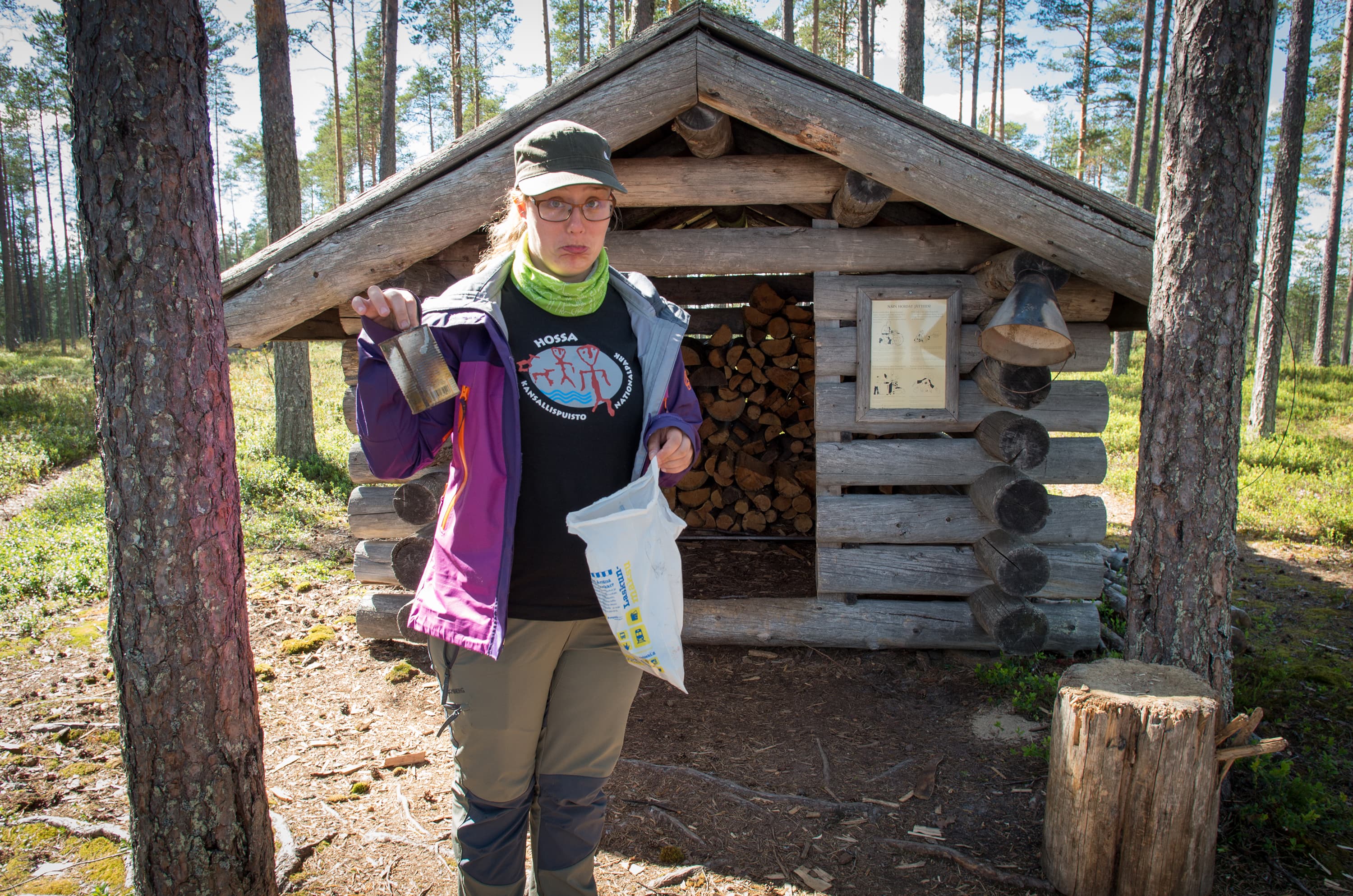 Image: Tuuli Turunen A woman stands in front of a log cabin with low ceiling in the forest, her expression displeased, holding a trash bag in one hand and litter in the other.