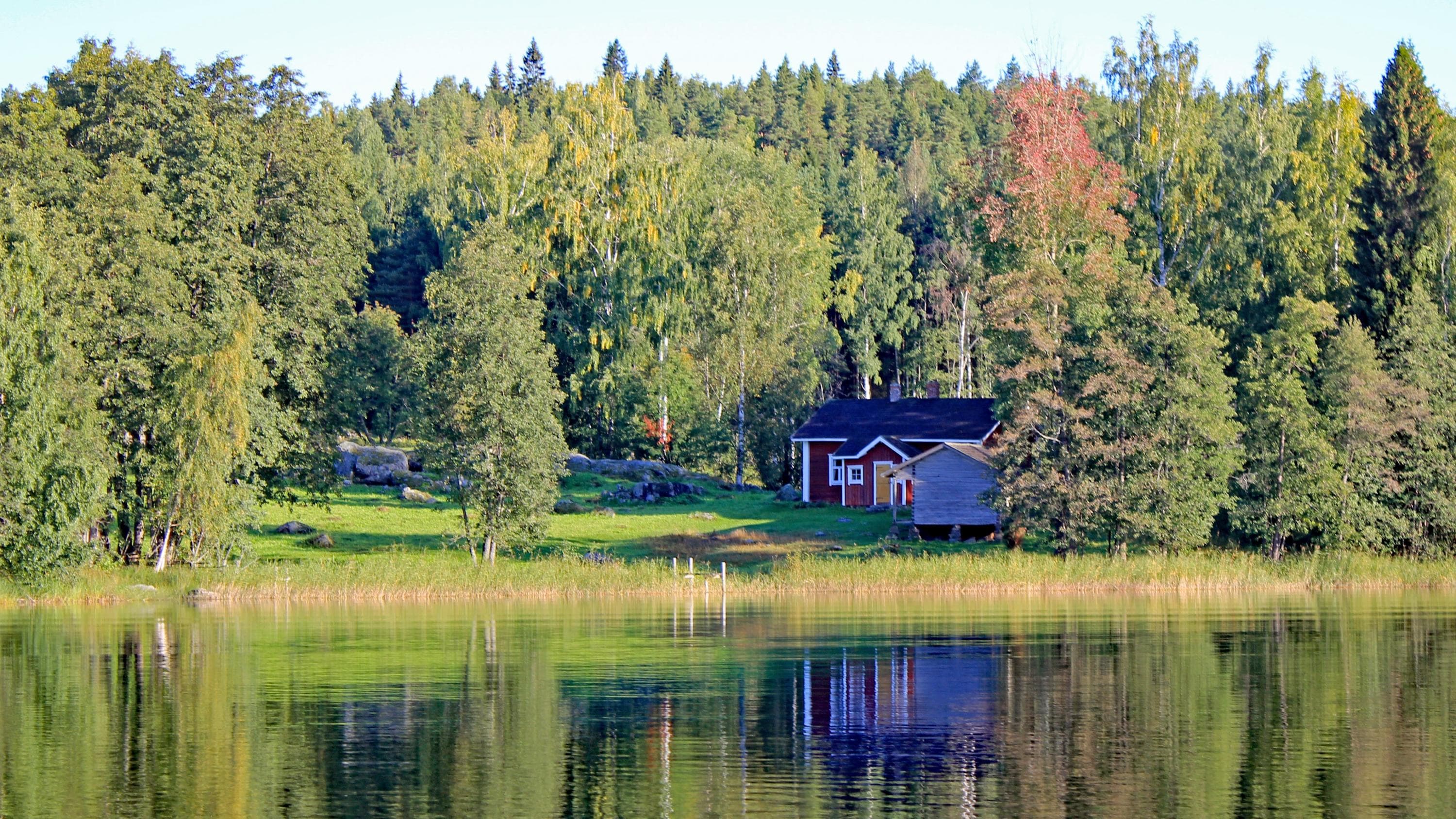 Image: Tarja Halme A landscape captured from the water. A calm water surface, an old croft by the shore, with forest in the background.