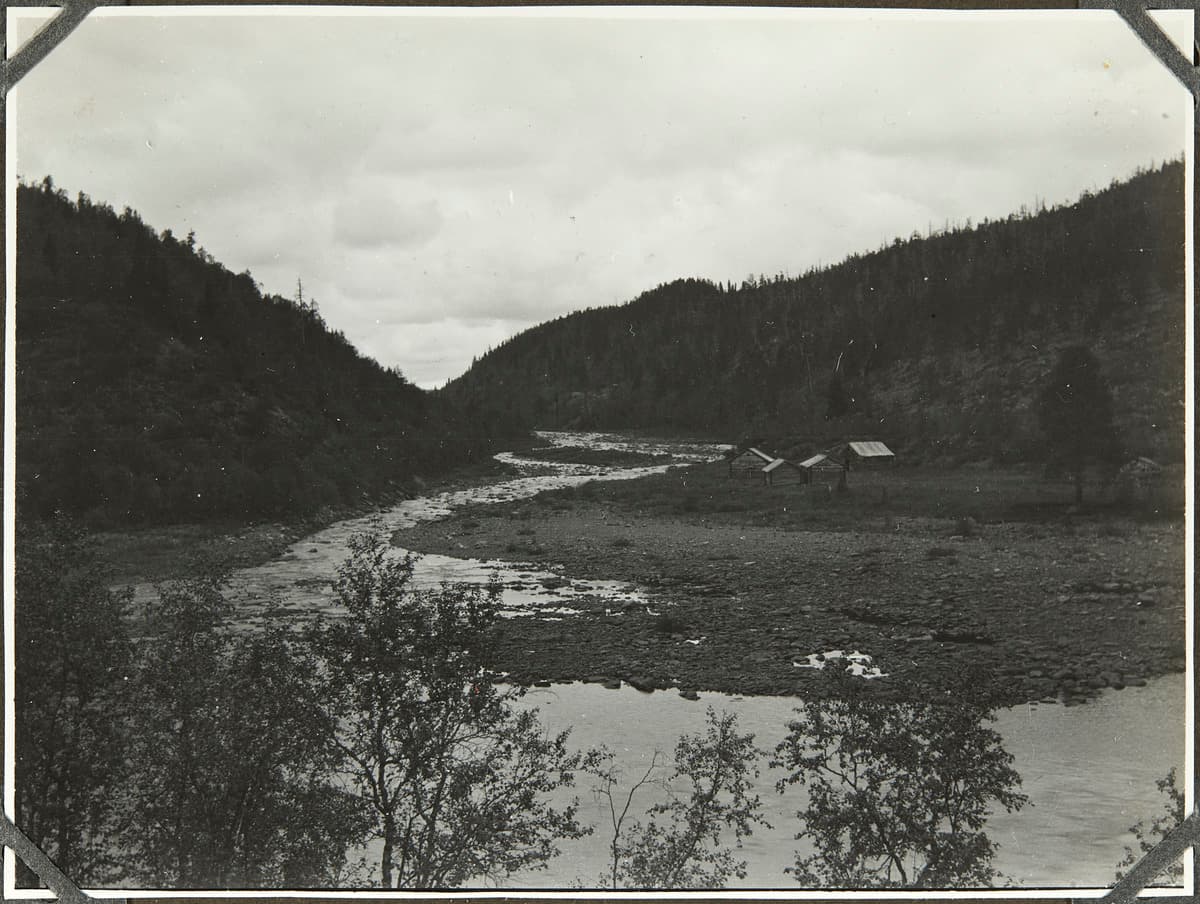 Image: Mikkola Erkki / Museovirasto, kansatieteen kuvakokoelma. A smaller river flows between the hills into a larger river. On the shore, there is a meadow and buildings.