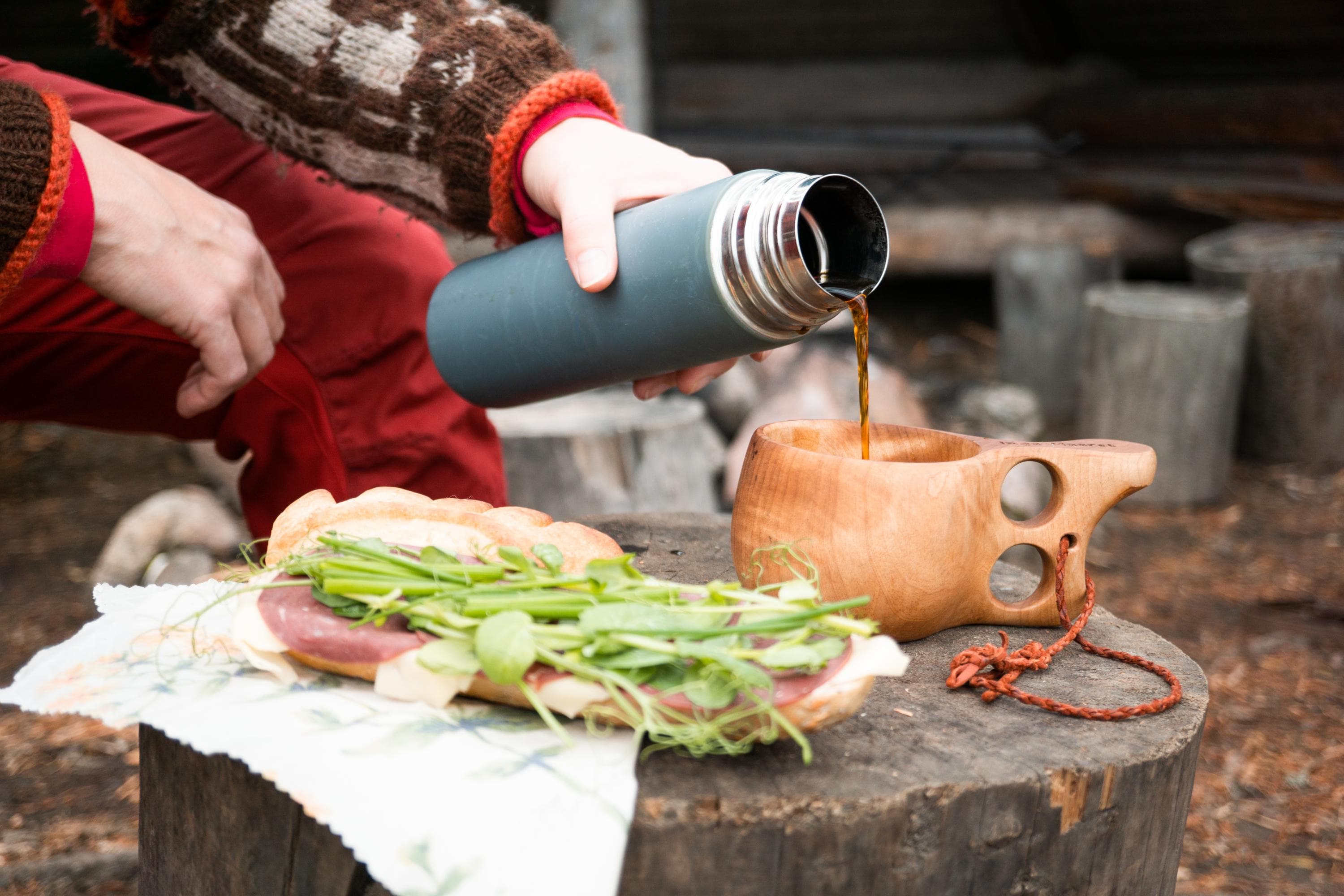 Image: Inga-Maaret Aikioniemi Sandwiches and a wooden cup (kuksa) are laid out on a tree stump. A hand is pouring coffee from a thermos into the kuksa.