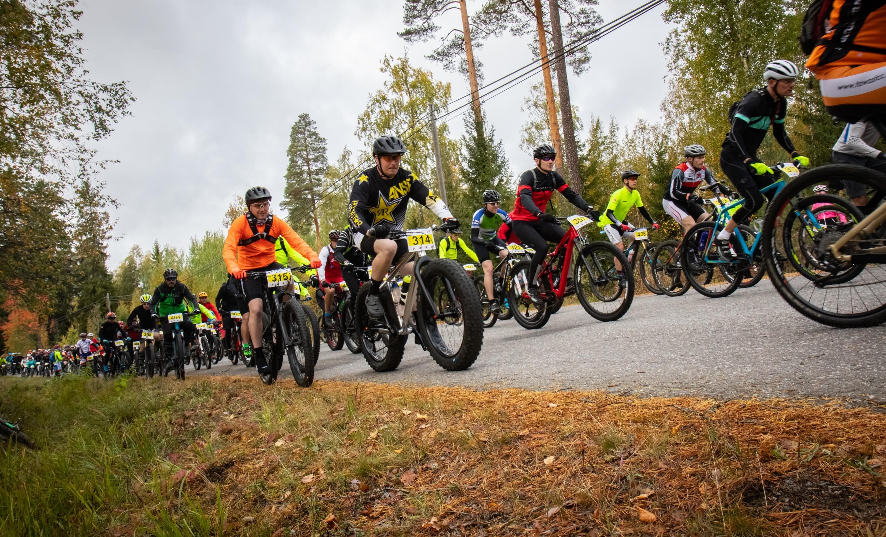 Kuva: Oikea Reitti Oy Kymmeniä värikkäisiin kisa-asuihin pukeutuneita maastopyöräilijöitä lähtevät matkaan asfalttitietä pitkin.
