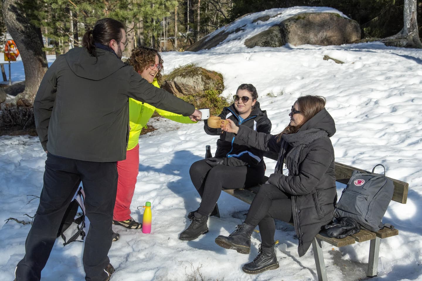 Image: VisitTampere Four people are toasting. The people are smiling, and two of them are sitting on a bench. There is snow on the ground, and the sun is shining.