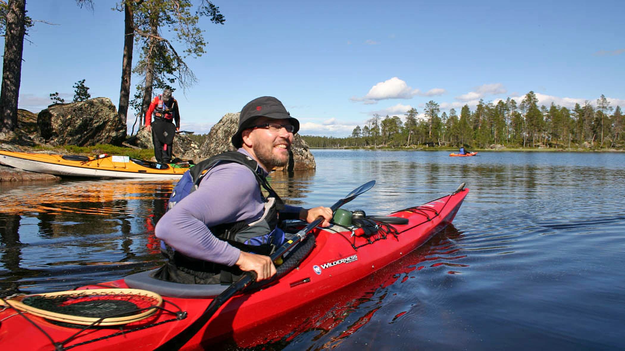 Image: Olli Vainio A paddler is sitting in a kayak, wearing a hat. The kayak is near the shore, and a hiker is standing on the beach. It is a sunny summer day.