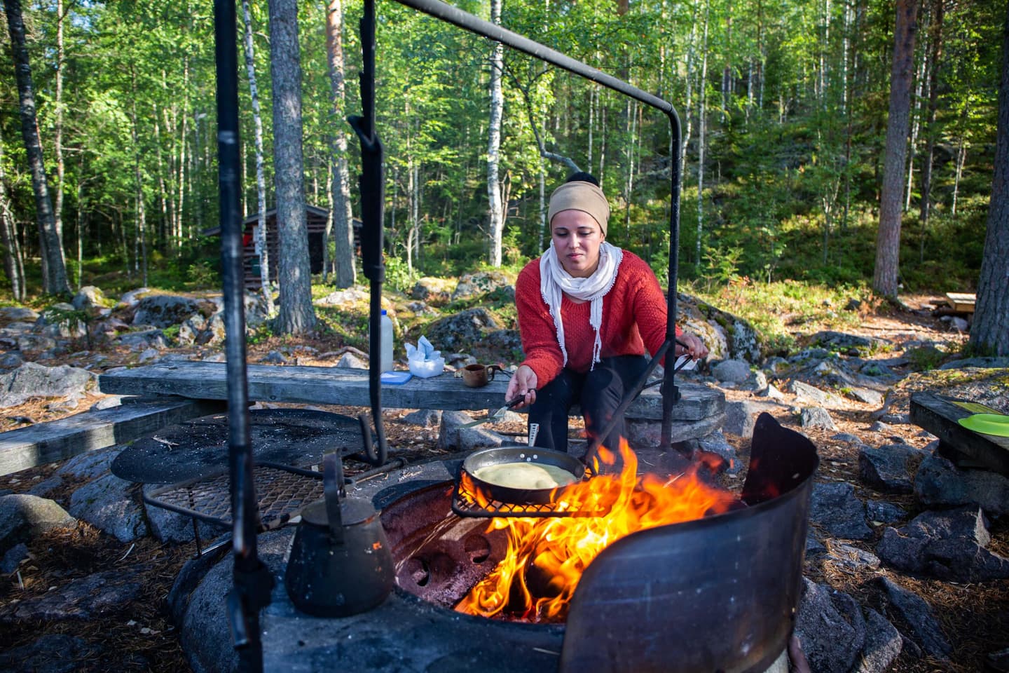 Image: Saara Lavi A hiker is frying pancakes at a campfire site. The fireplace has a grate and various types of hooks that can be used to assist with cooking.