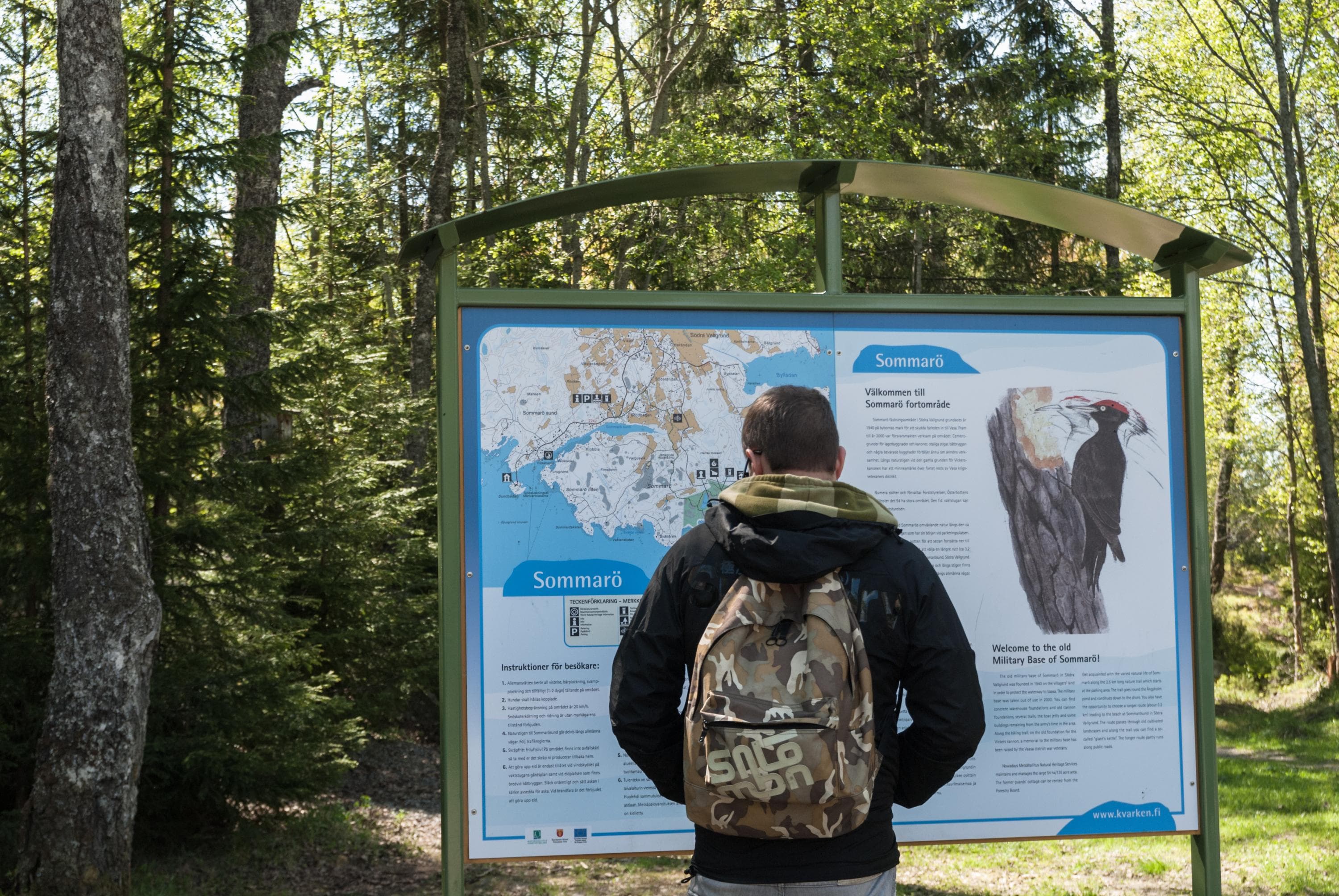 Kuva: Tuija Waren Poika tarkastelee aurinkoisena päivänä luontopolkutaulun karttaa päiväreppu selässä, taustalla kuusimetsää.
