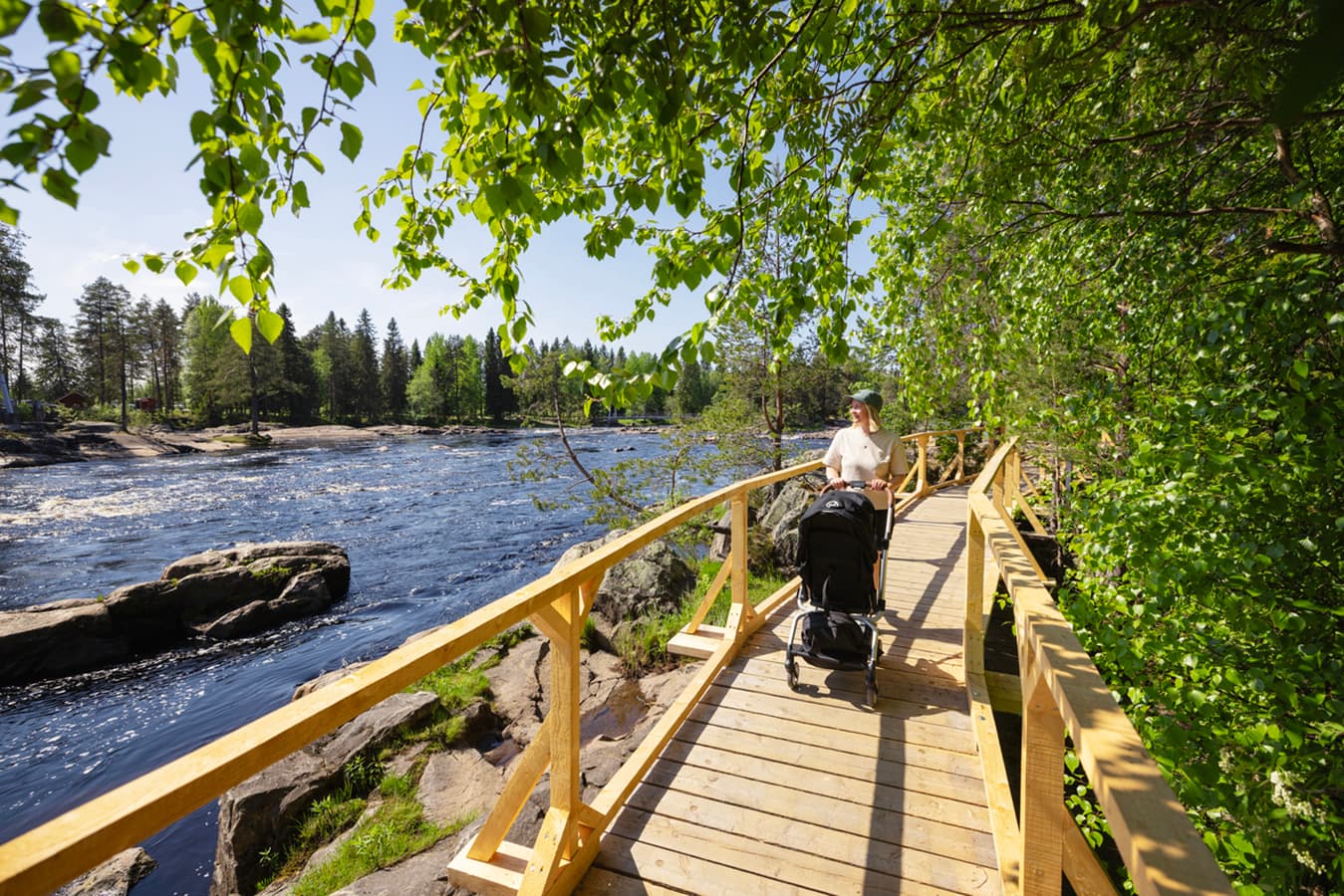 Image: Oulun kaupunki / Harri Tarvainen A hiker is pushing a pram on a wide boardwalk by the river. There are railings on both sides of the path.