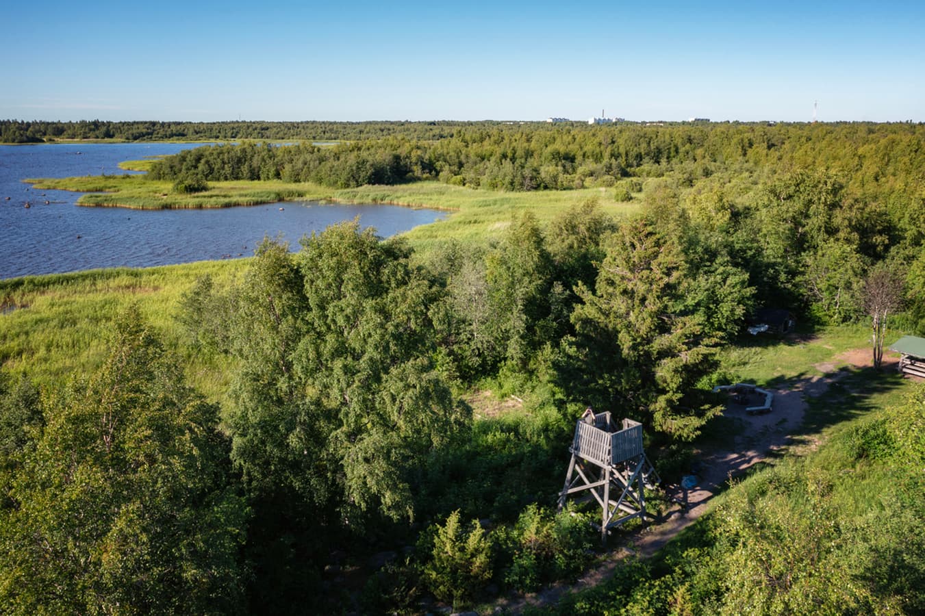 Image: Oulun kaupunki / Harri Tarvainen Aerial view of a birdwatching tower near the shore, where a hiker is looking at the sky through binoculars. Next to the tower, there is a campfire area.