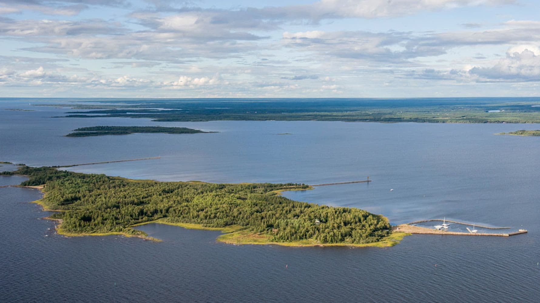 Image: Lentokuva Vallas Oy An aerial view of a sea island. At the southern tip of the island, there are sailing ships docked in the harbor. Most of the island is covered by lush forest. In the background, the mainland is visible.