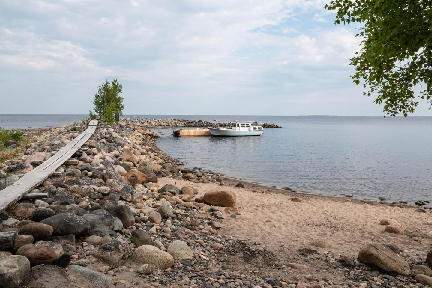 Image: Ismo Lampi The white boat is moored to the pier. Wooden duckboards lead from the pier to the island. On the island's shore, there is sand and stones.