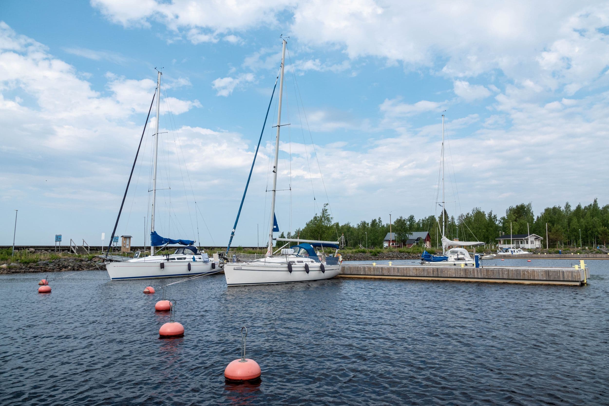 Image: Ismo Lampi Three sailboats are moored to a long pier. On the island behind the pier, there are three buildings and a deciduous forest.
