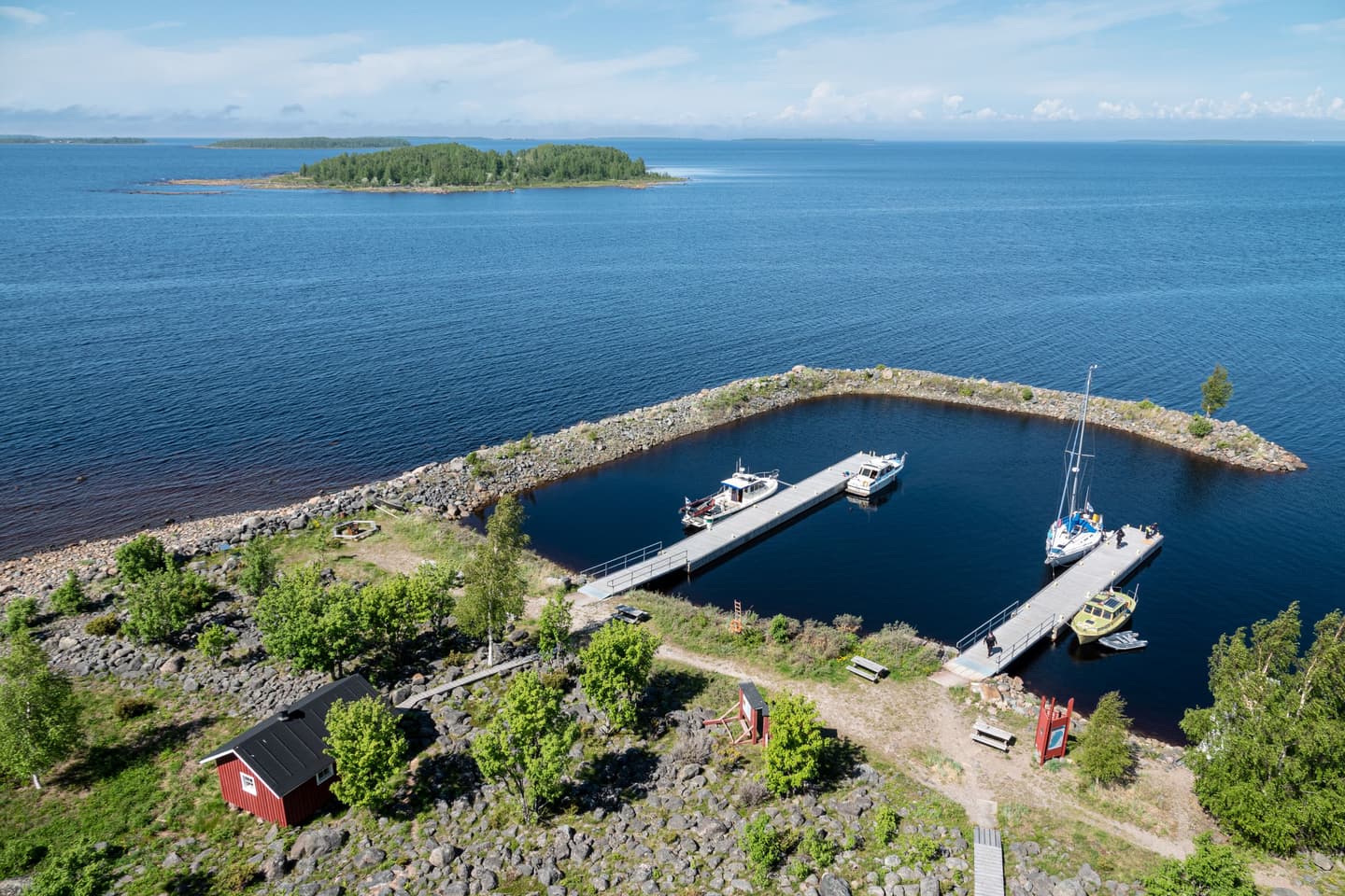 Image: Ismo Lampi The harbor basin seen from above. There are two long piers in the harbor, with boats moored alongside. On the island’s shore, there is a small red building surrounded by lush vegetation.