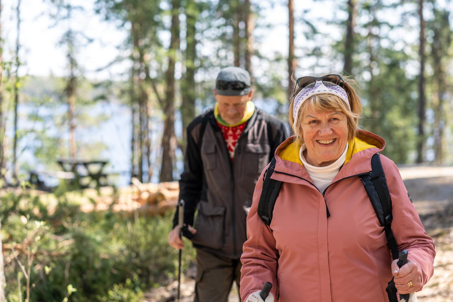 Image: Mitja Piipponen Two smiling hikers with walking poles. Behind them are trees with glimpses of water between the trunks.