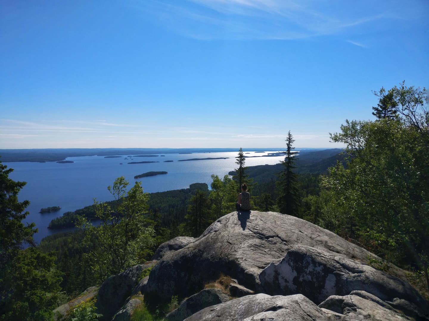 Image: Elina Ode A hiker sits on a high cliff in summer, looking out over the lake landscape below.