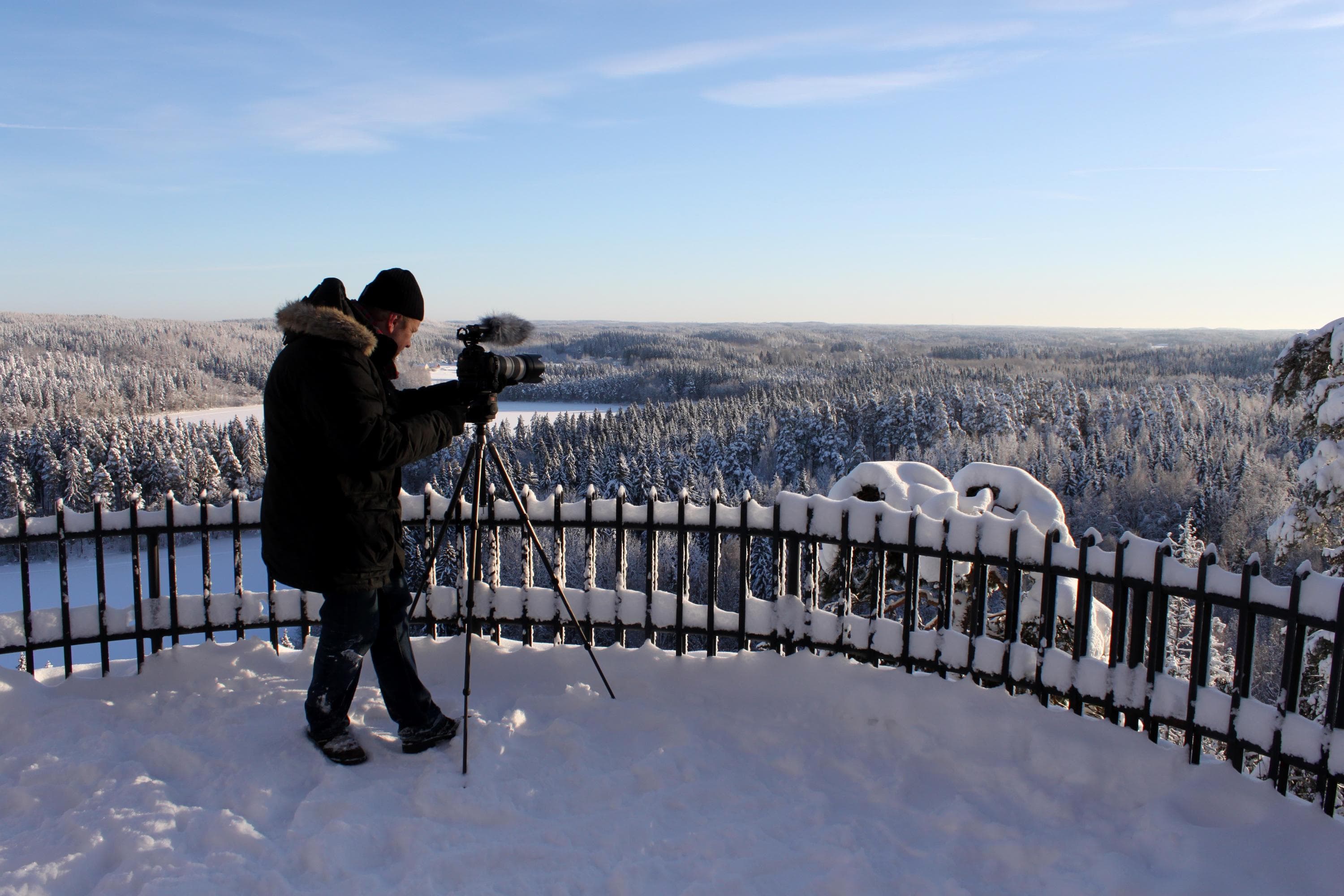 Kuva: S-M Hiukkamäki Retkeilijä näköalatasanteella kameran ja kolmijalan kanssa. Kamera on suunnattu kohti lumista metsää.