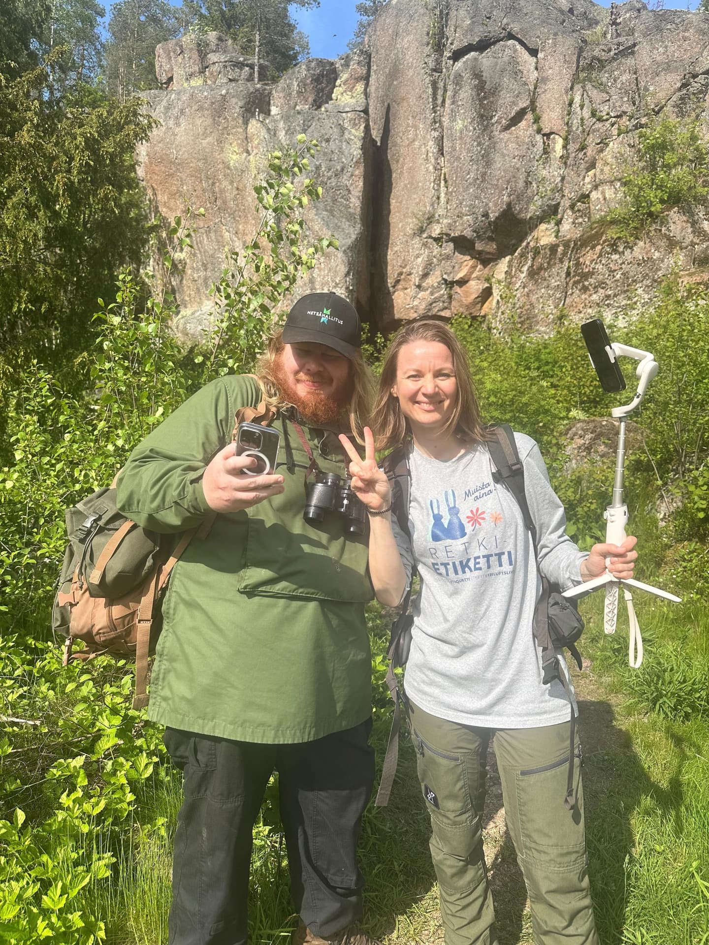 Kuva: Kirsi Kallio Kaksi henkilöä poseeraa kuvaajalle kallioseinämän edessä kesällä. Toisella on kädessään kännykkä ja kaulassa kiikarit ja toisella on kädessään selfiekeppi ja päällään Retkietiketti-paita.