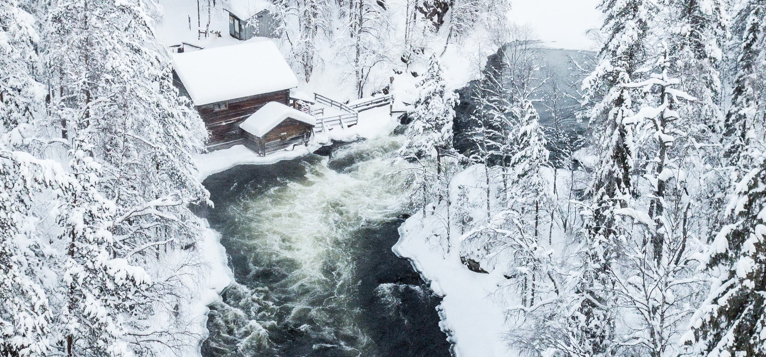 Bild: Kahtelis Oy Ett vinterlandskap i Oulanka nationalpark. I mitten forsar ett strömt vatten och en bro, omgiven av snötäckt skog.