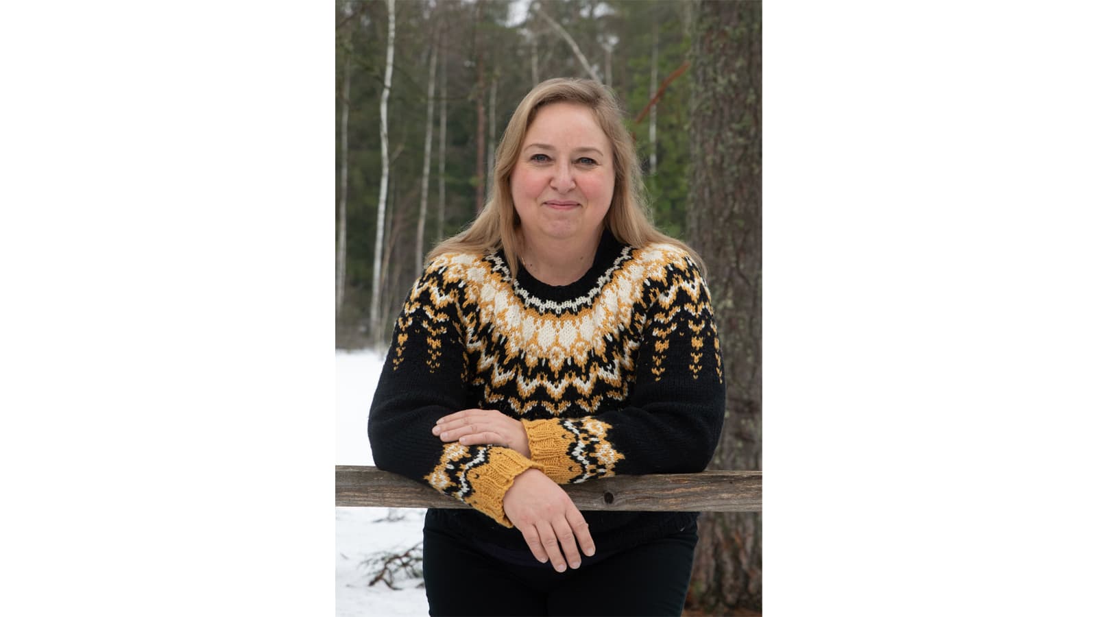 Image: Tanja Seppälä A smiling person leans against a wooden railing in winter. The person is wearing a black wool jumper with orange, white, and black patterns on the upper part and cuffs.