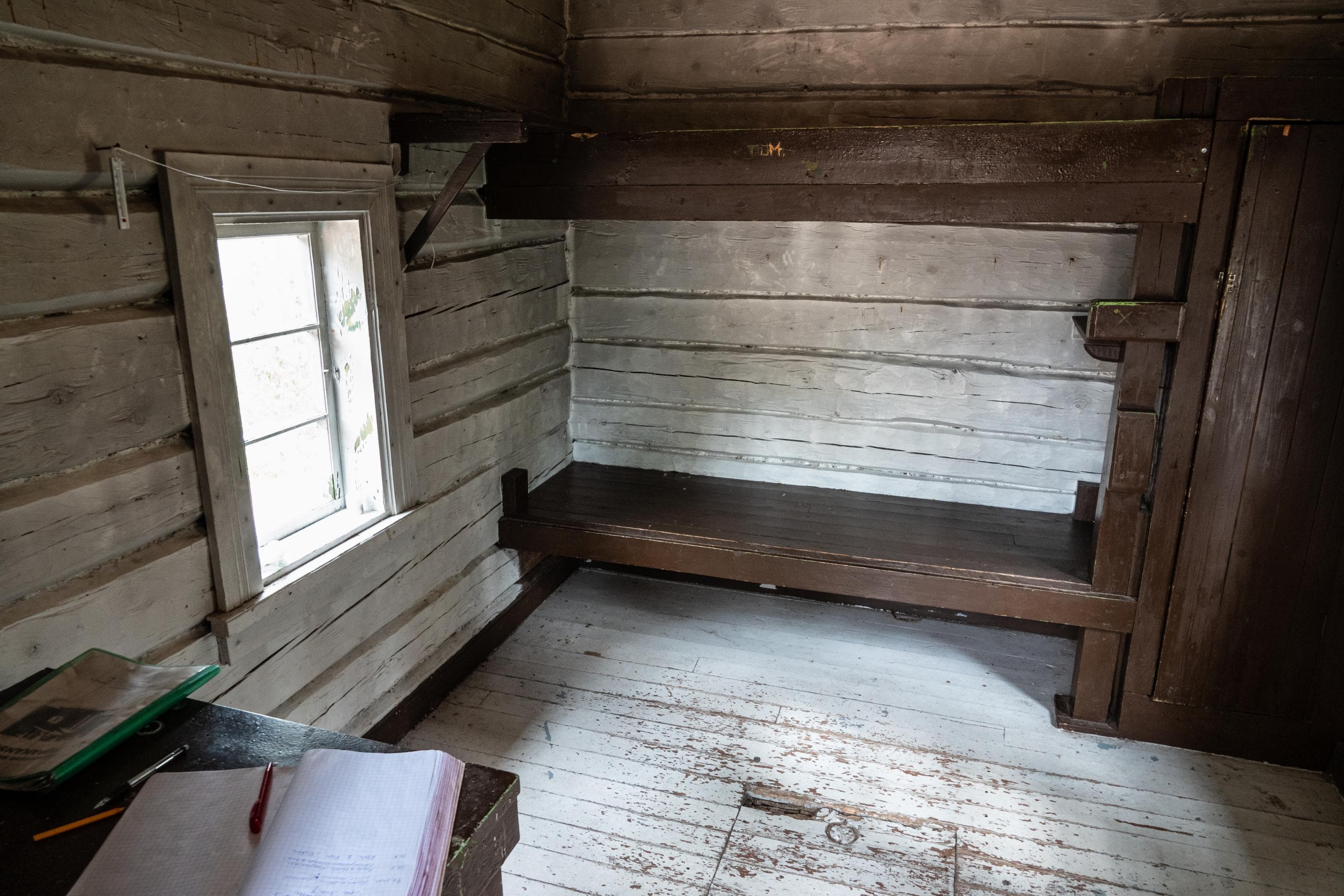 Kuva: Ismo Lampi Pieni valkoiseksi maalattu hirsimökki sisältä. Takaseinällä puinen kerrossänky. Vasemmalla seinällä ikkuna. Etualalla pöytä, jossa on vieraskirja.