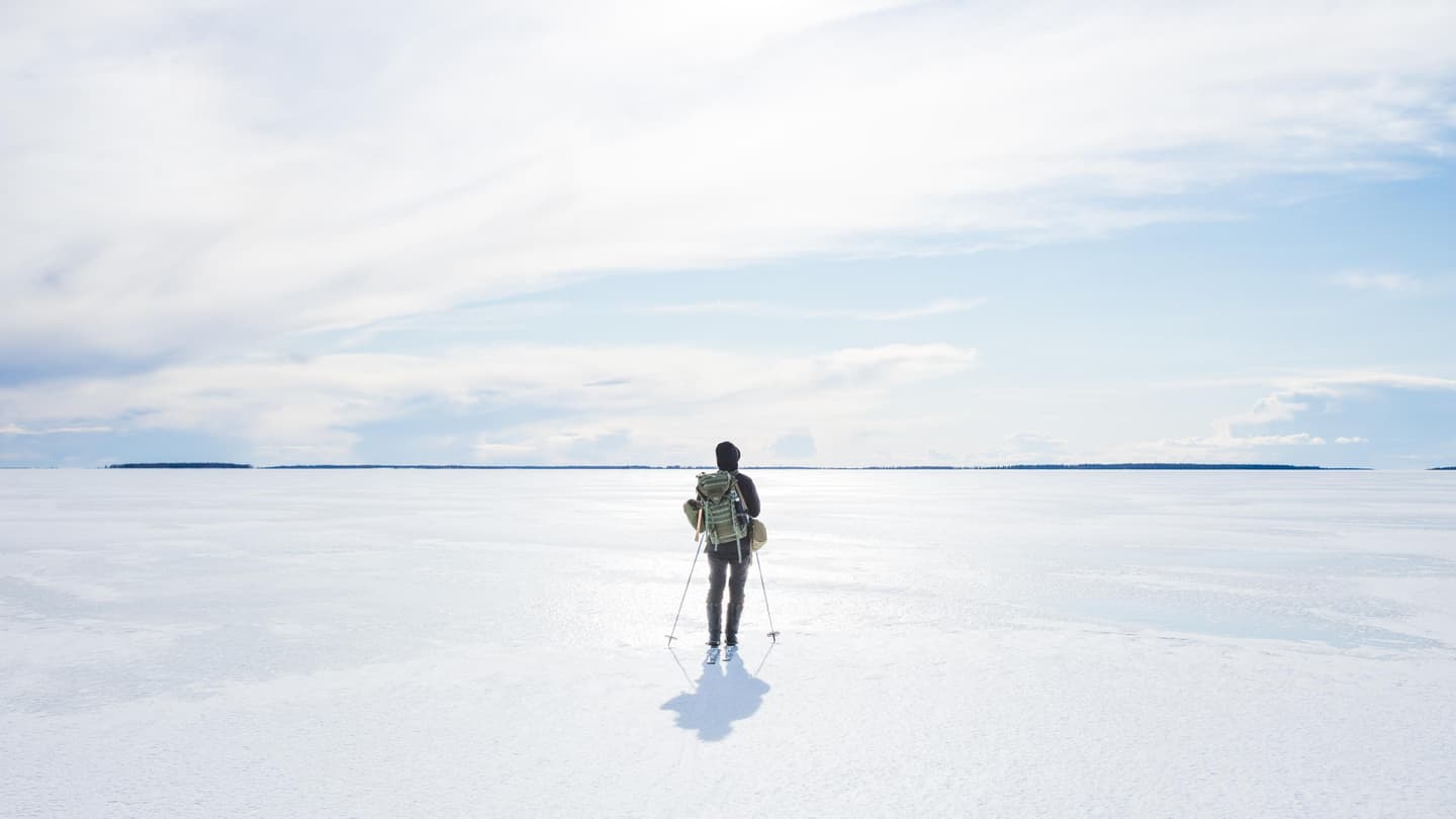 Kuva: Jarno Hamalainen Hiihtäjä rinkkansa kanssa valkoisella jäällä. Aurinko paistaa kirkkaasti pilviverhon takaa.
