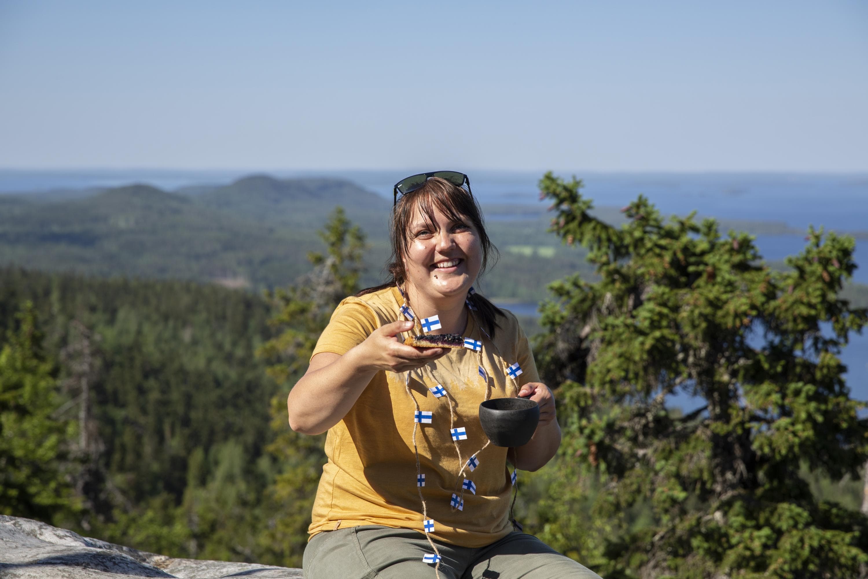 Image: Saara Lavi A hiker holds a piece of blueberry pie in one hand and a kuksa cup in the other. Around the hiker’s neck hangs a ribbon garland made of small Finnish flags. The hiker sits on rocky terrain with a scenic view of hills and waters in the background.