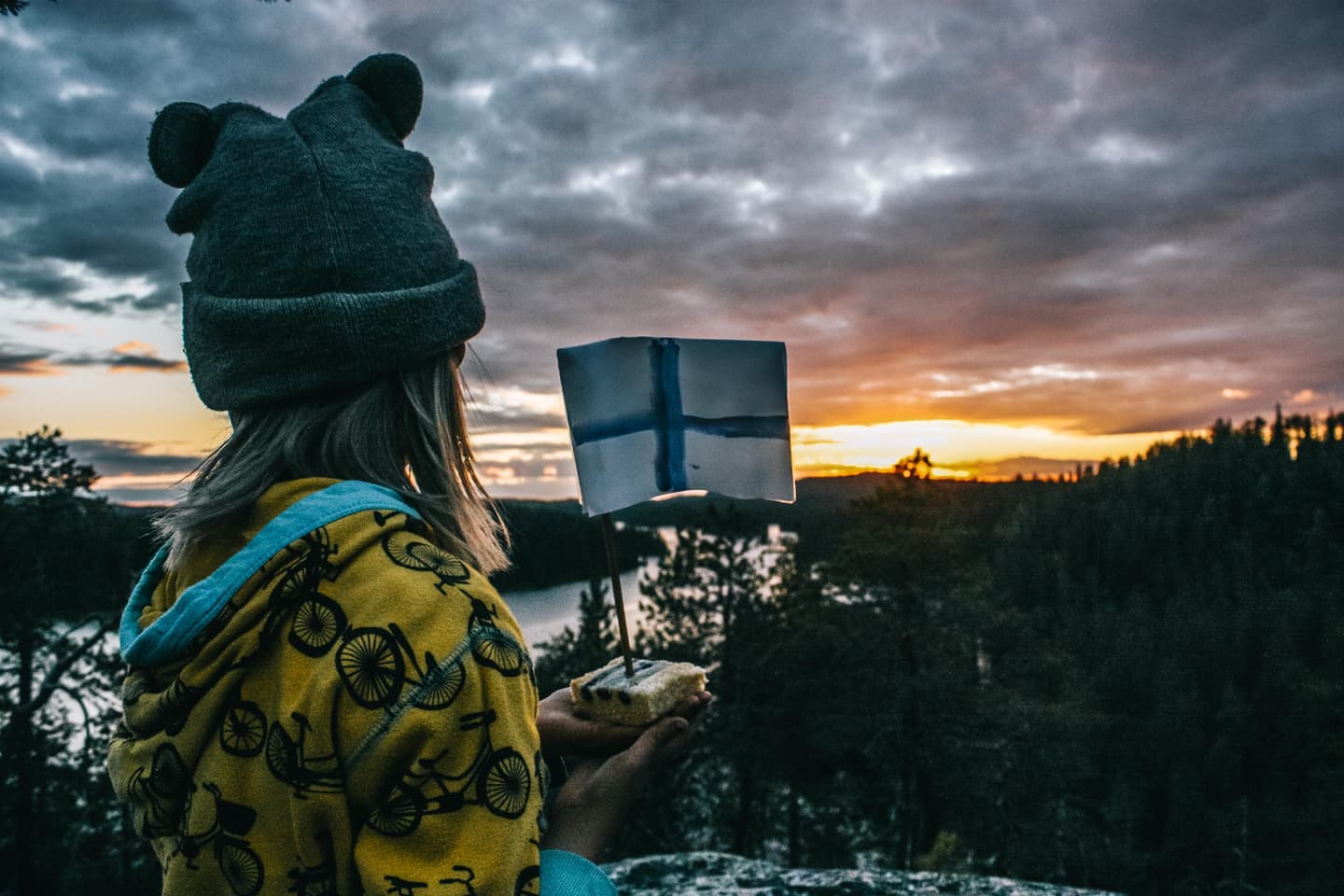 Image: Marjo Kämärainen In the foreground, a child holds a piece of blueberry pie with a small Finnish flag standing upright in it. The child is looking away from the camera towards the sunset. Forest and water are visible in the background.