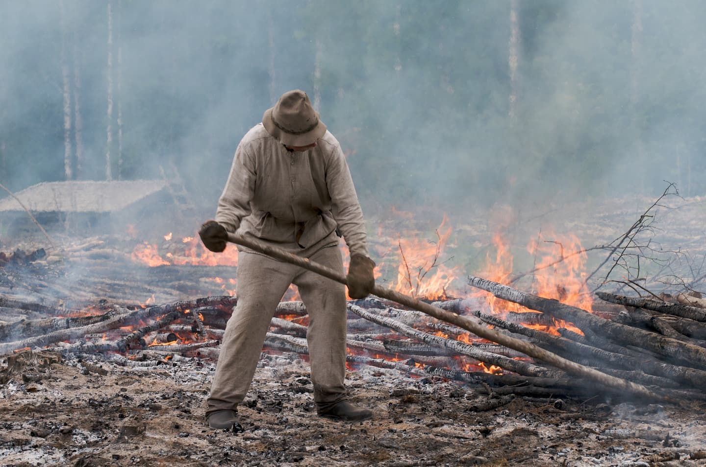 Image: Jouni Borgman A person in traditional clothing shifts the burning soil forward using special tools as part of the slash-and-burn process. Through the smoke, a building and the forest edge can be seen in the background.