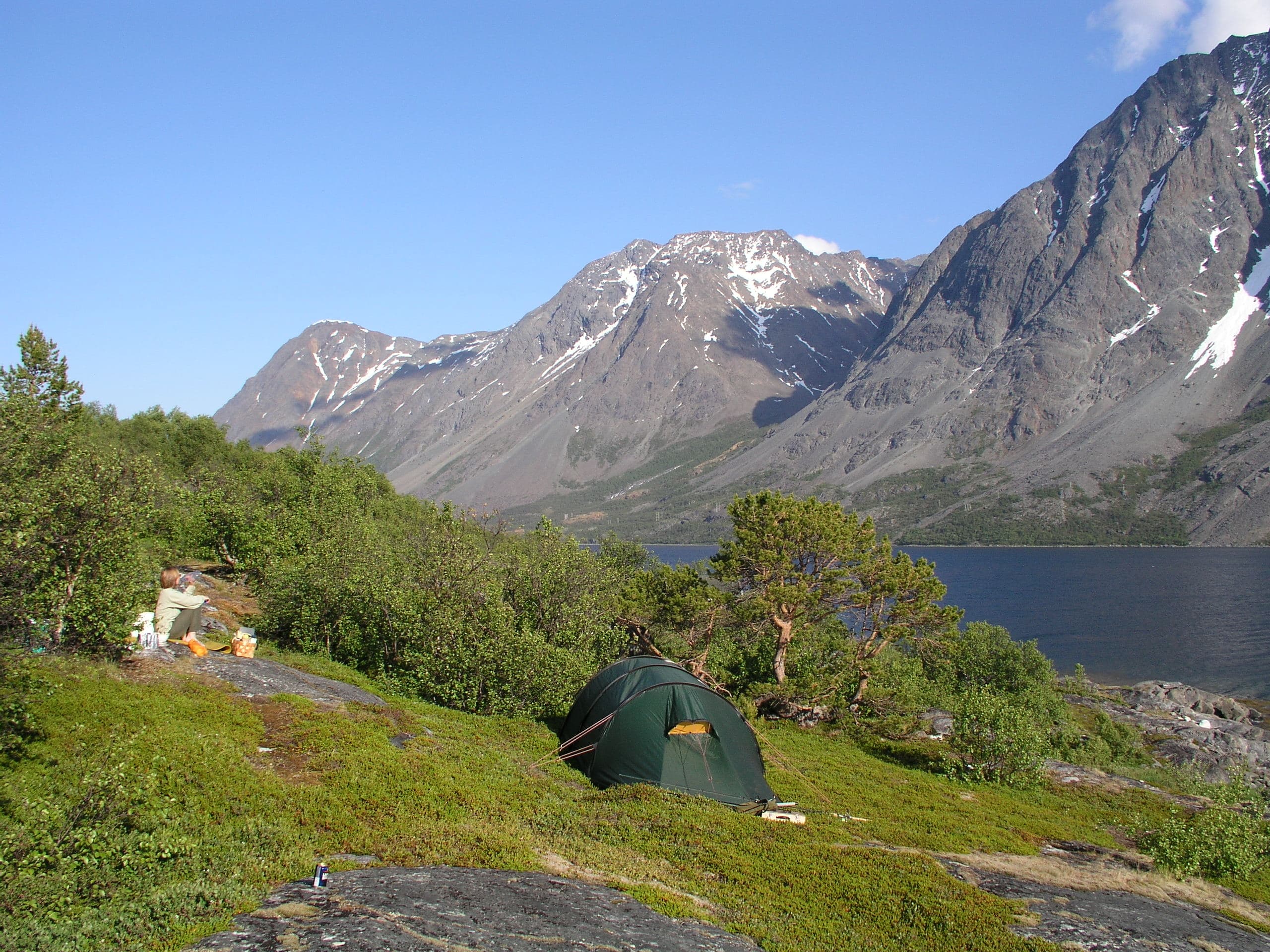 Kuva: Ari Karhilahti Vuoristomaisema vuonon rannalta. Etualalla pusikkoon pystytetty teltta ja retkeilijä tauolla.