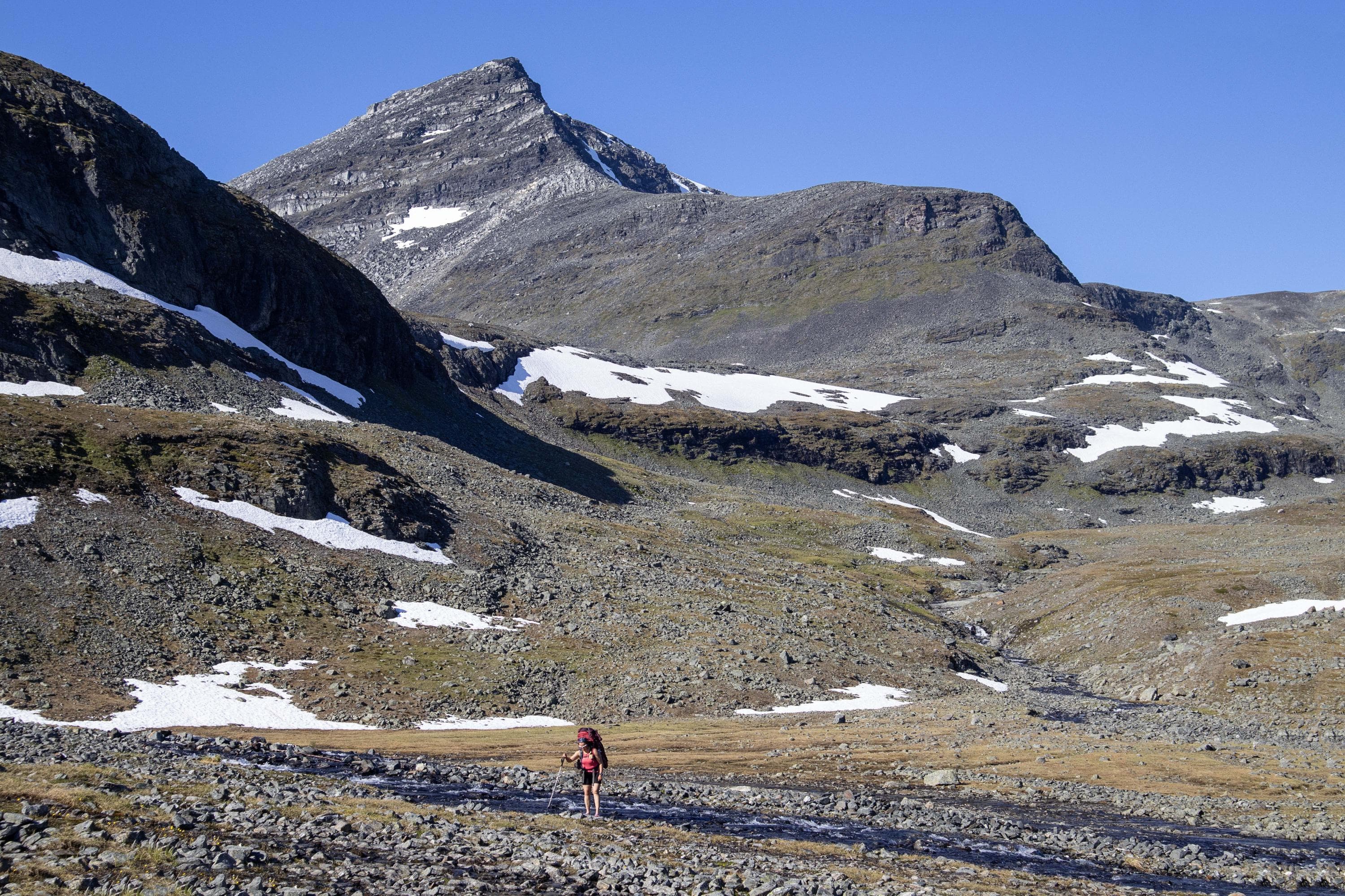 Kuva: Anna Pakkanen Tunturin juurella joki. Vaeltaja rinkka selässään ja sauva kädessään seisoo avojaloin rannalla selin jokeen.