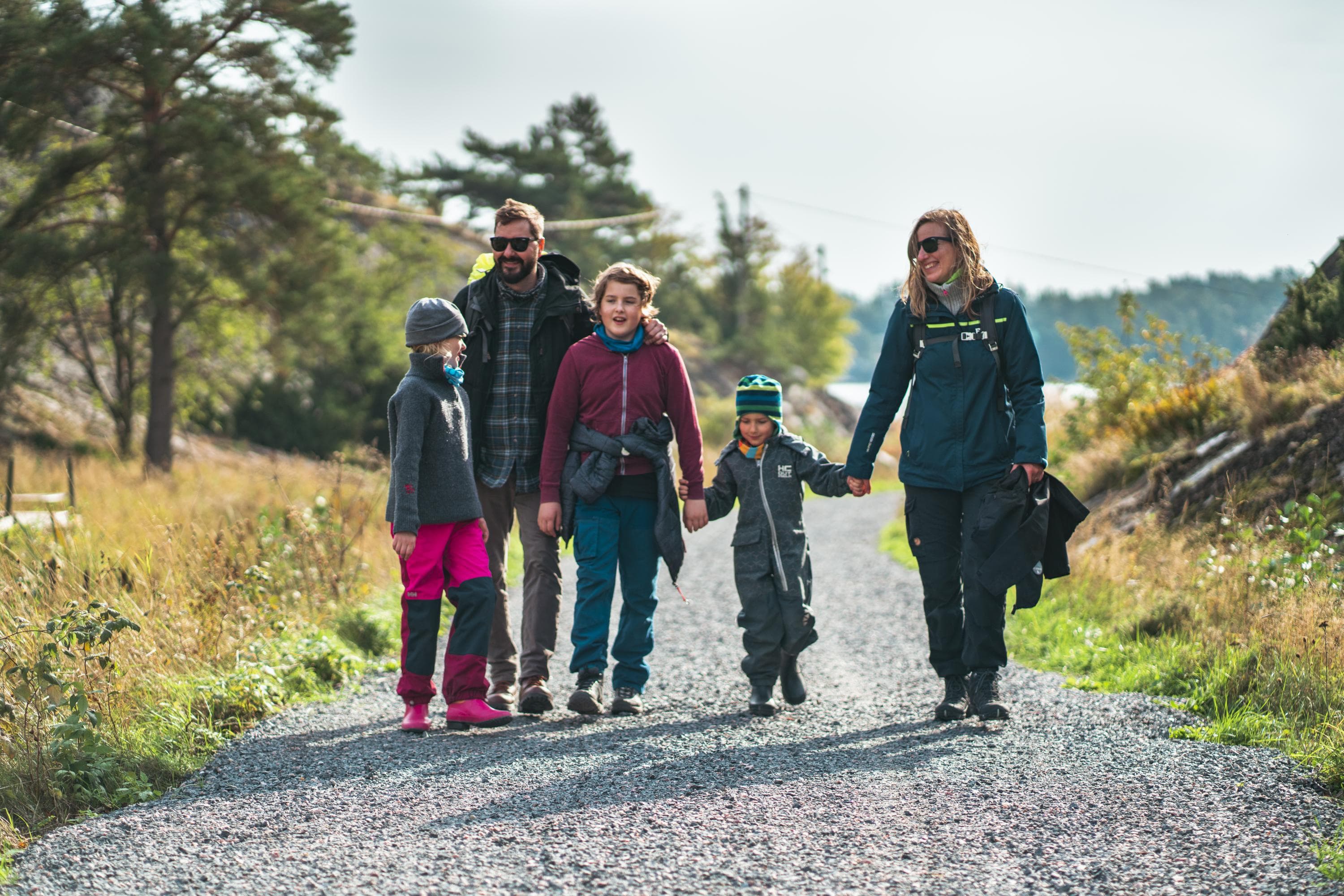Kuva: One Take Productions Kaksi aikuista ja kolme lasta kävelemässä hiekkatiellä kävelemässä syksyisessä maisemassa. Takana häämöttää vettä ja vastaranta. Osa ihmisistä pitää toisiaan kädestä kiinni.