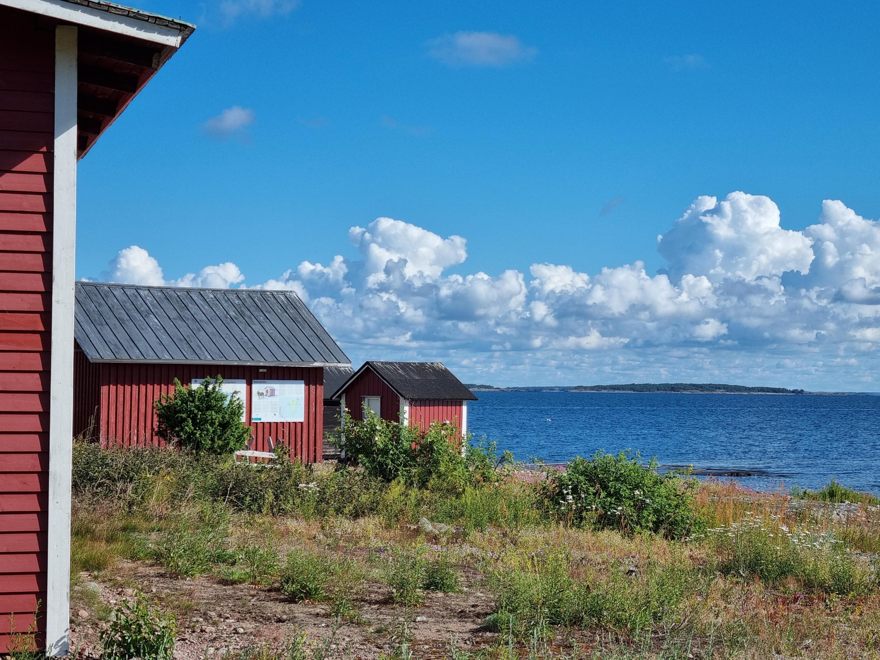 Kuva: Marja Hokkanen Punaisia puutaloja rannalla, taustalla sininen meri ja kirkas taivas.