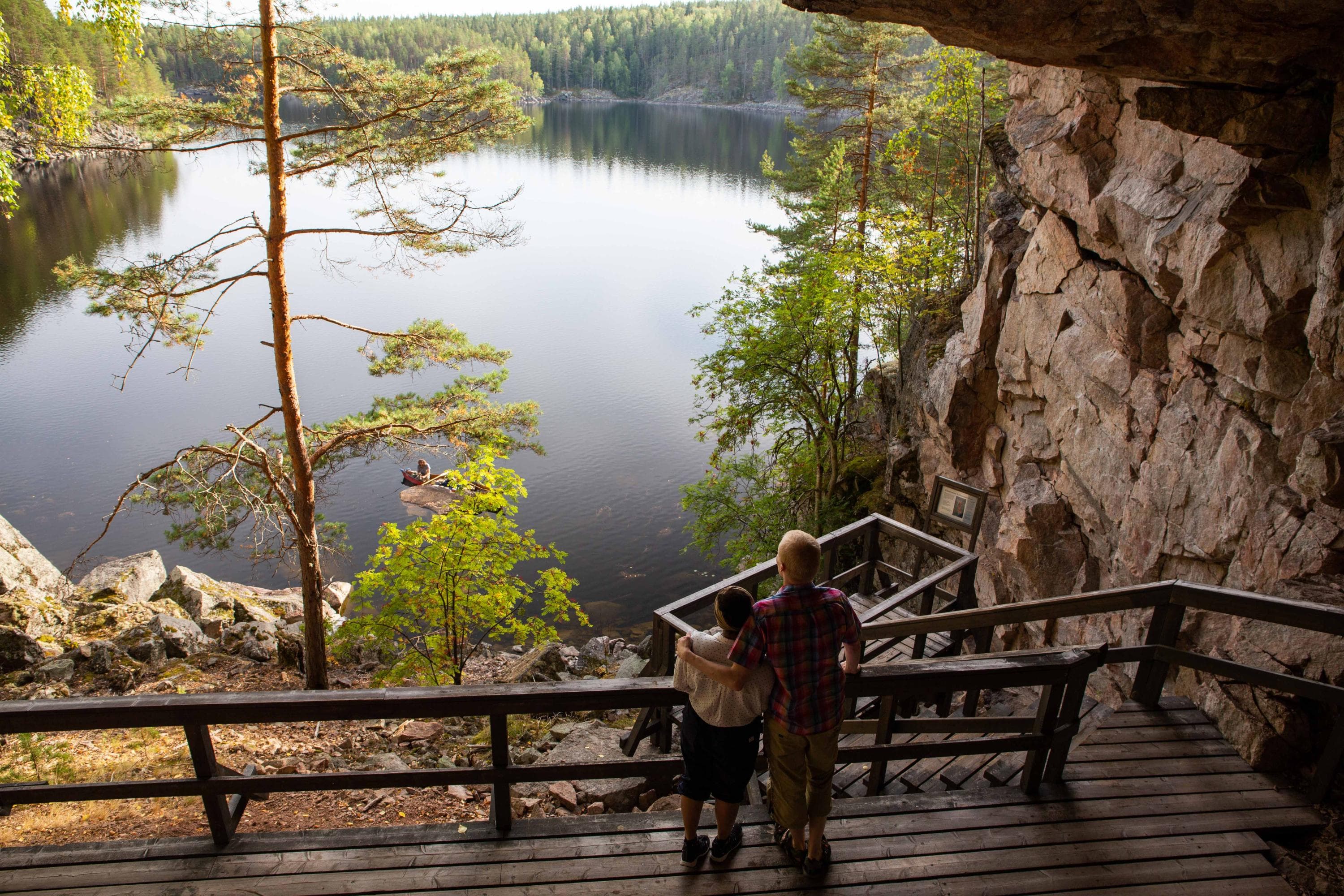Kuva: Saara Lavi Kaksi retkeilijää luolassa näköalatasanteella. Taustalla peilityyni kesäinen järvimaisema.