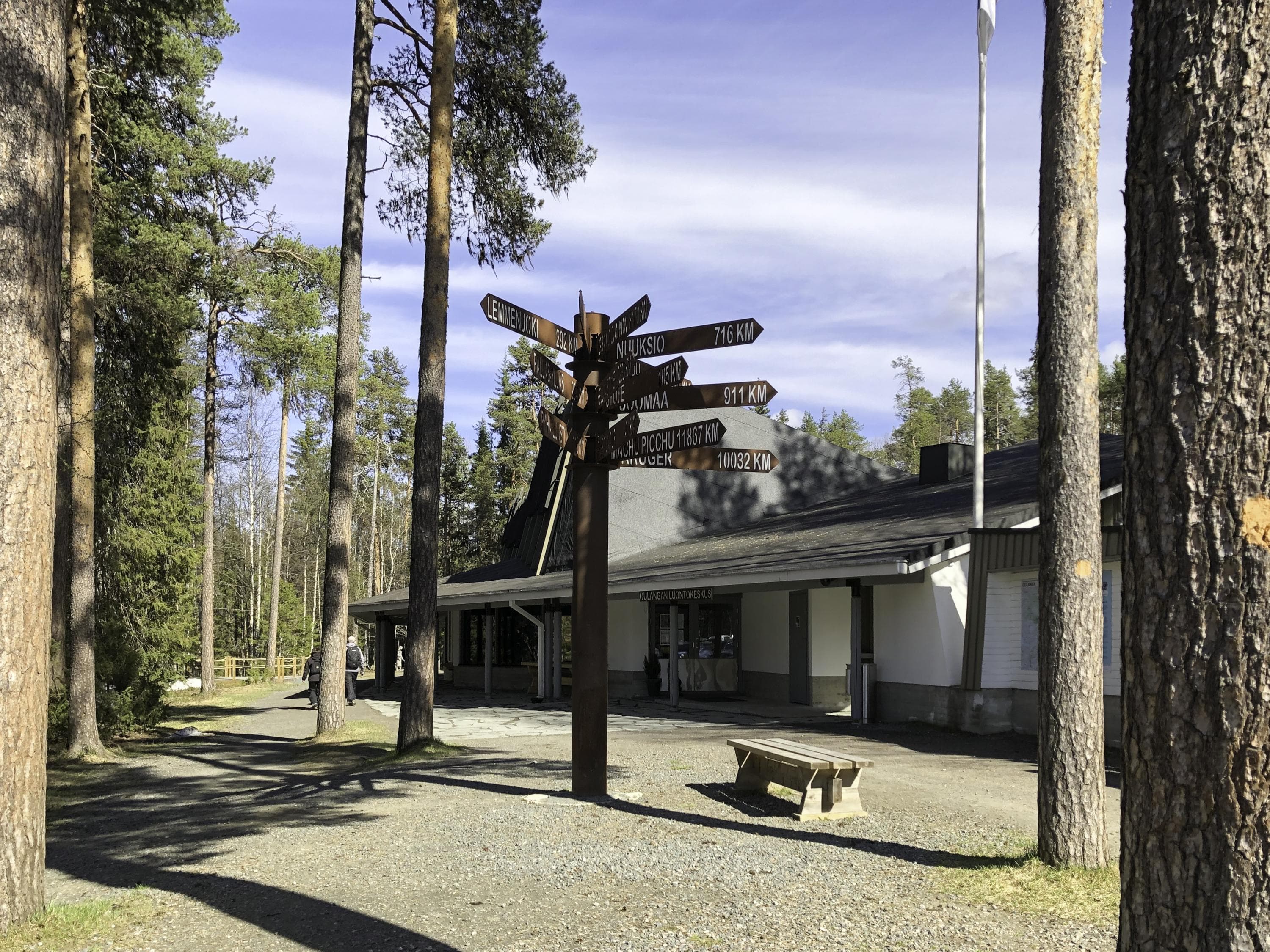 Kuva: Minna Niinistö Vaalean rakennuksen sisäänkäynnin edustalla on liuskekivilaatta. Piha on hiekkaa ja siinä kasvaa suuria mäntyjä. Etualalla on pylväs, jossa on paljon opasteviittoja.