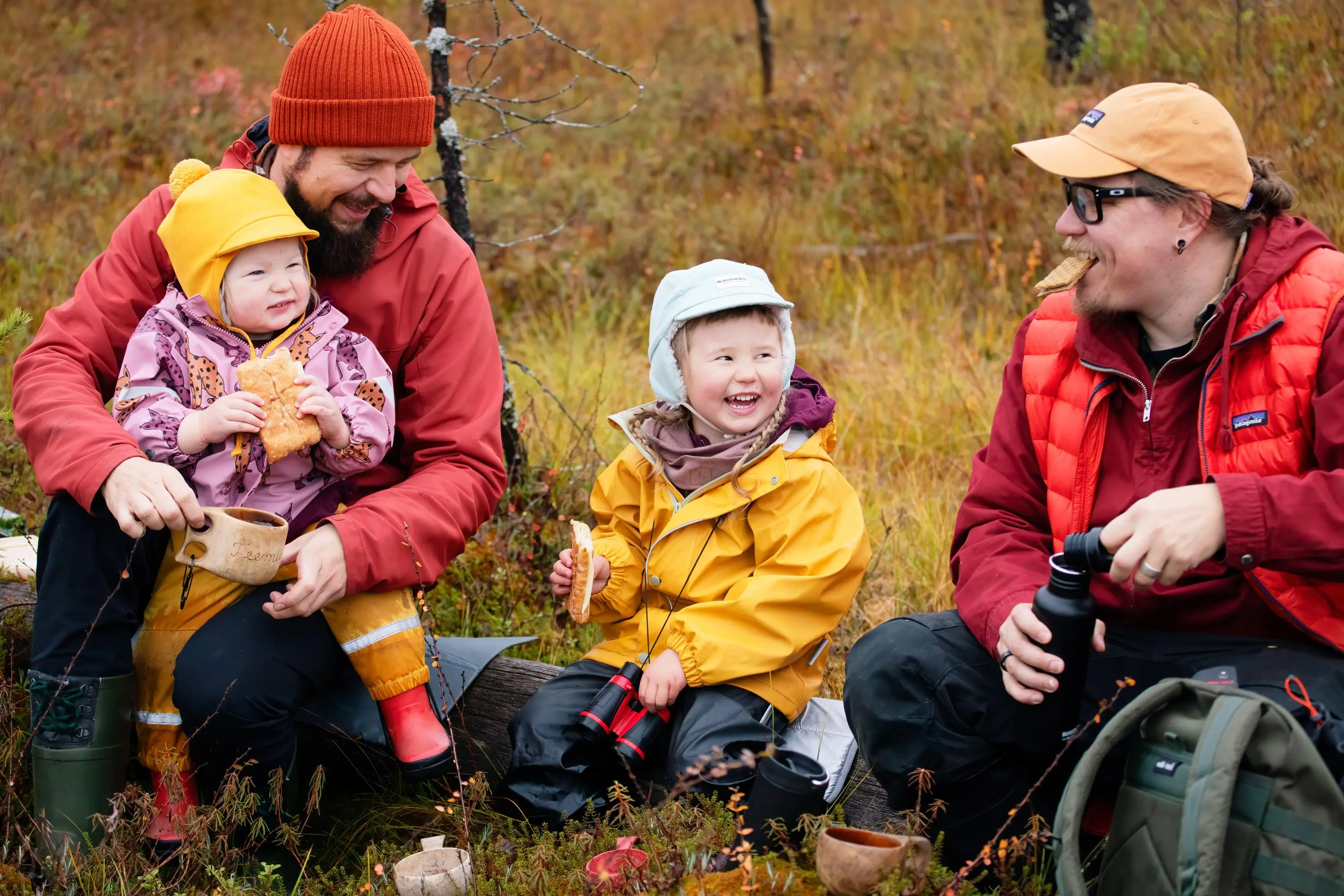 Kuva: Katri Lehtola / Keksi Kaksi aikuista punaisissa takeissa ja kaksi sadeasuista lasta istuu kaatuneella puulla syksyisellä suolla. Käsissä eväsleivät ja termospullo, vieressä eväsreppu.