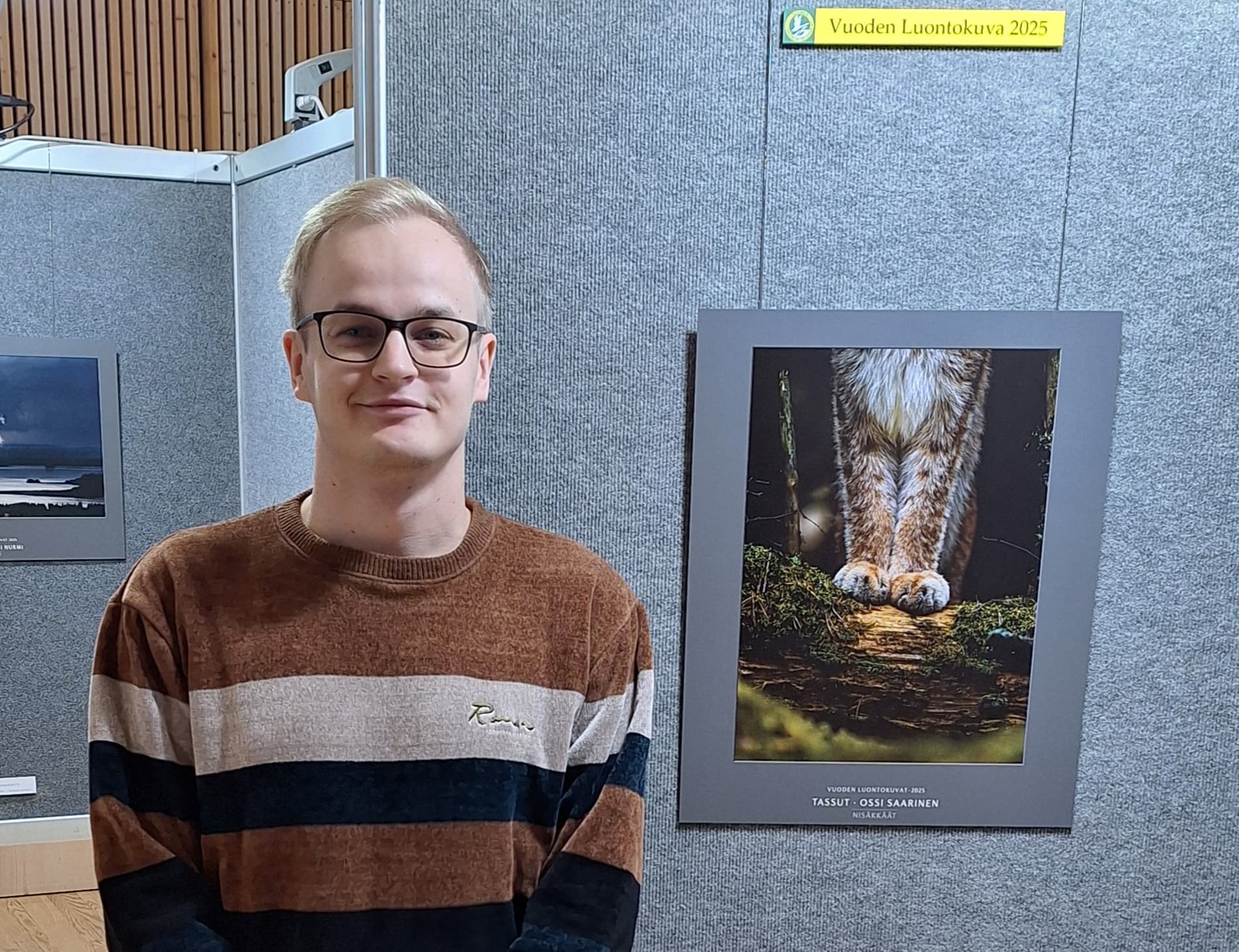 Kuva: Katja Määttä Ossi Saarinen seisoo näyttelytilassa. Saarisen takana on seinälle ripustettuna vuoden luontokuva 2025 ilveksen tassuista.