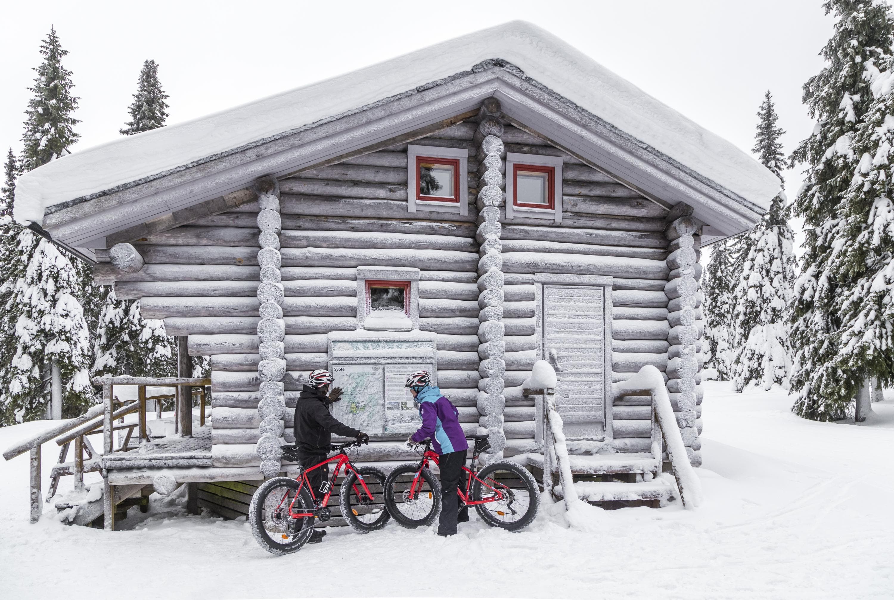 Kuva: Heikki Ketola / Vastavalo Kaksi pyöräilijää tutkimassa opastetauluja, jotka on kiinnitetty lumikuorrutetun hirsirakennuksen seinään. Tuvan ympärillä on kuusimetsää.