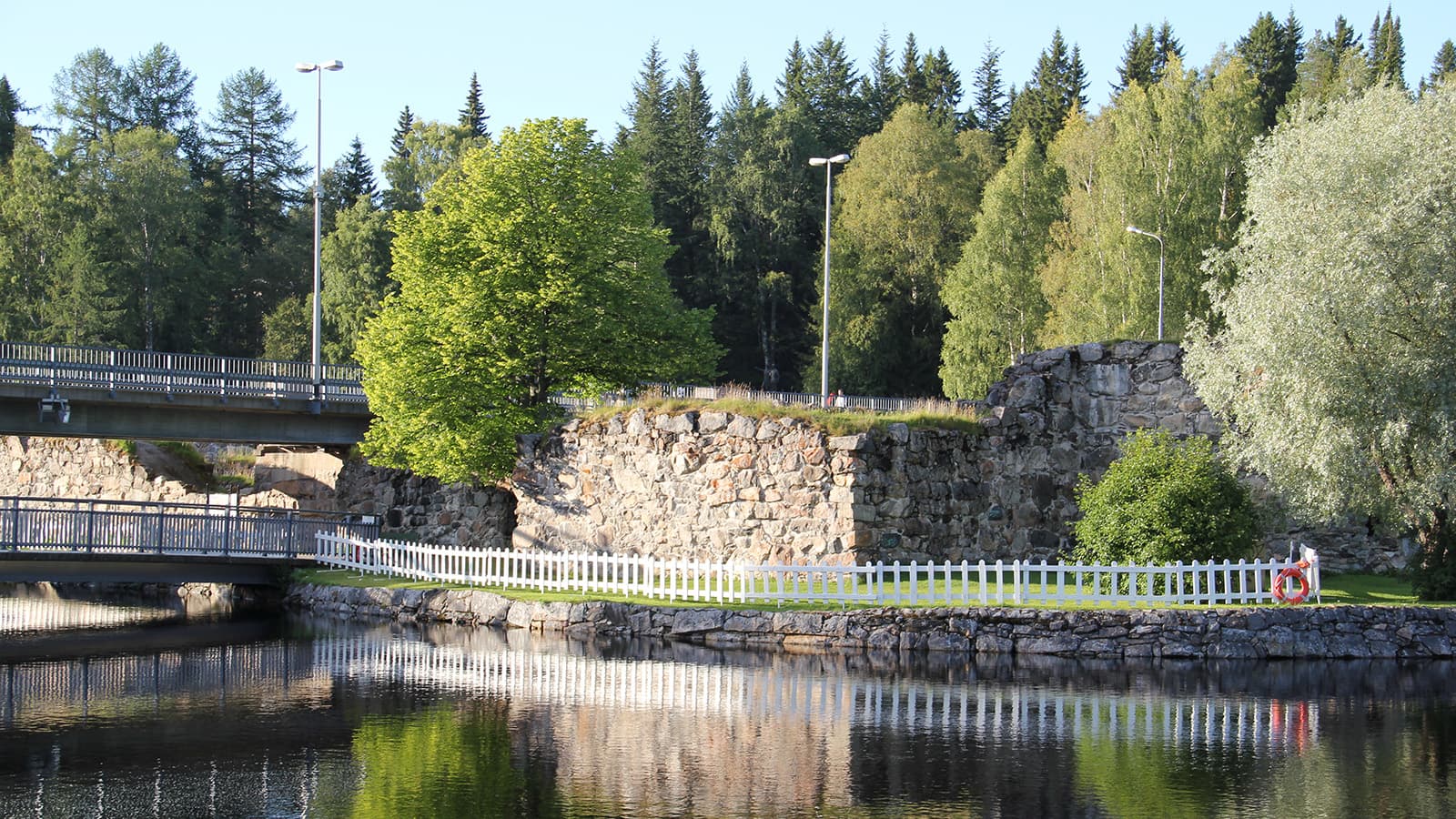 Kuva: Minna Niinistö Rauniolinna veden partaalla kesällä. Rantaa ja kulkuväylää on aidattu. Raunioiden ylitse kulkee tie.