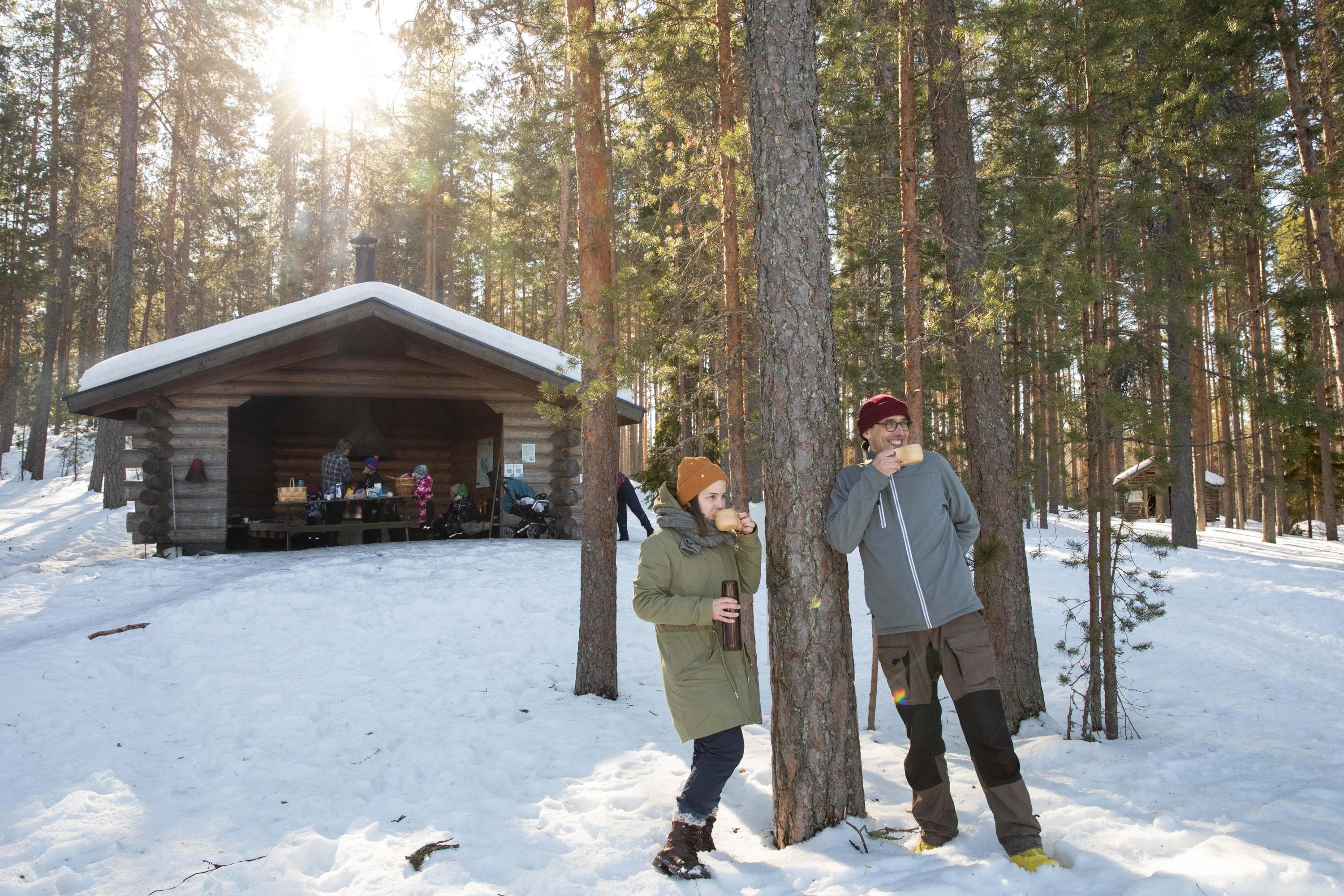 Bild: Mitja Piipponen Ett vintermotiv med två personer i förgrunden och fler personer som pausar med matsäck i ett vindskydd i bakgrunden.