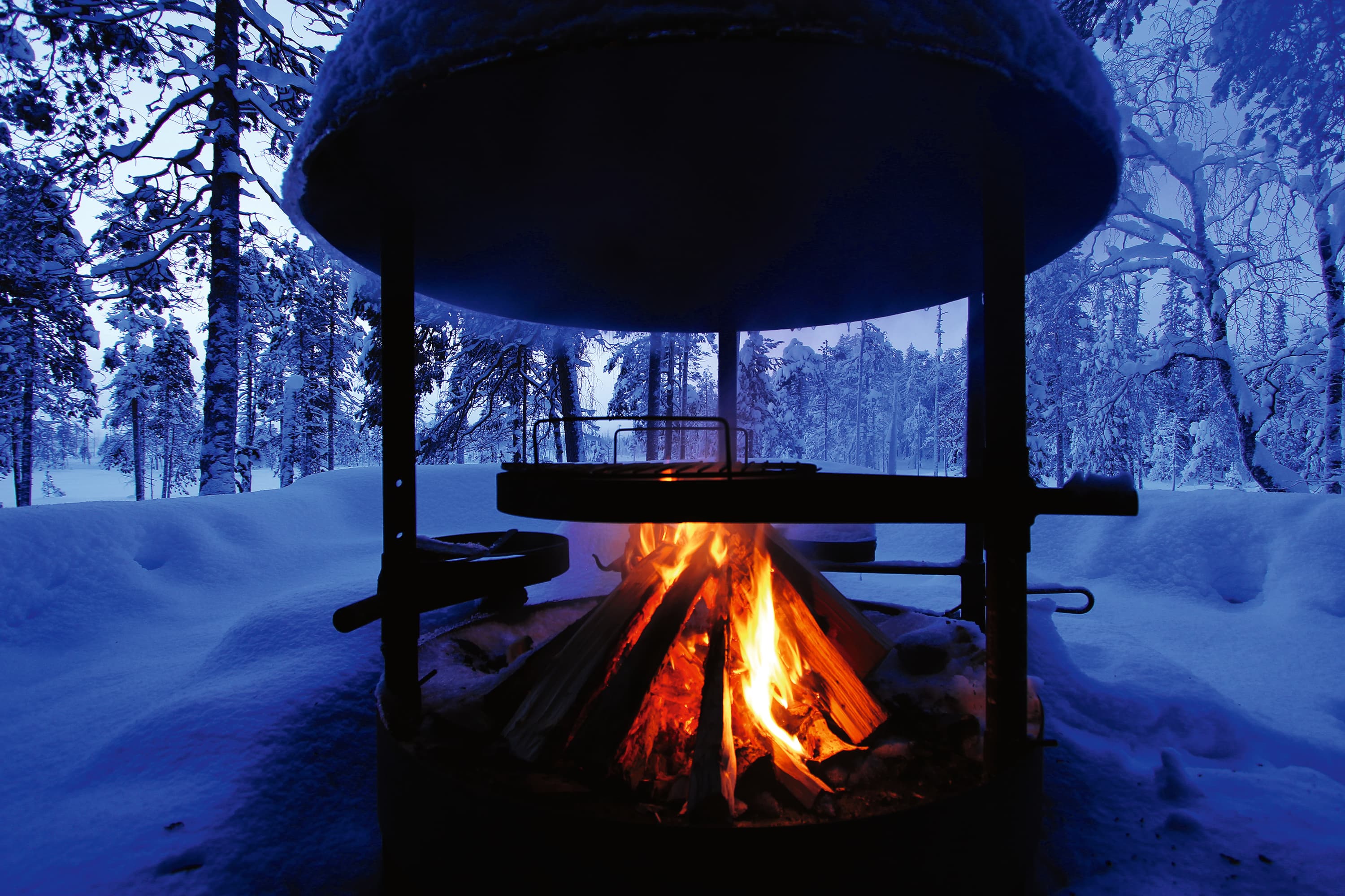 Kuva: Juha Paso Lumisen maiseman keskellä, sinertävässä hämärässä on hormillinen tulentekopaikka, jossa palaa tuli.