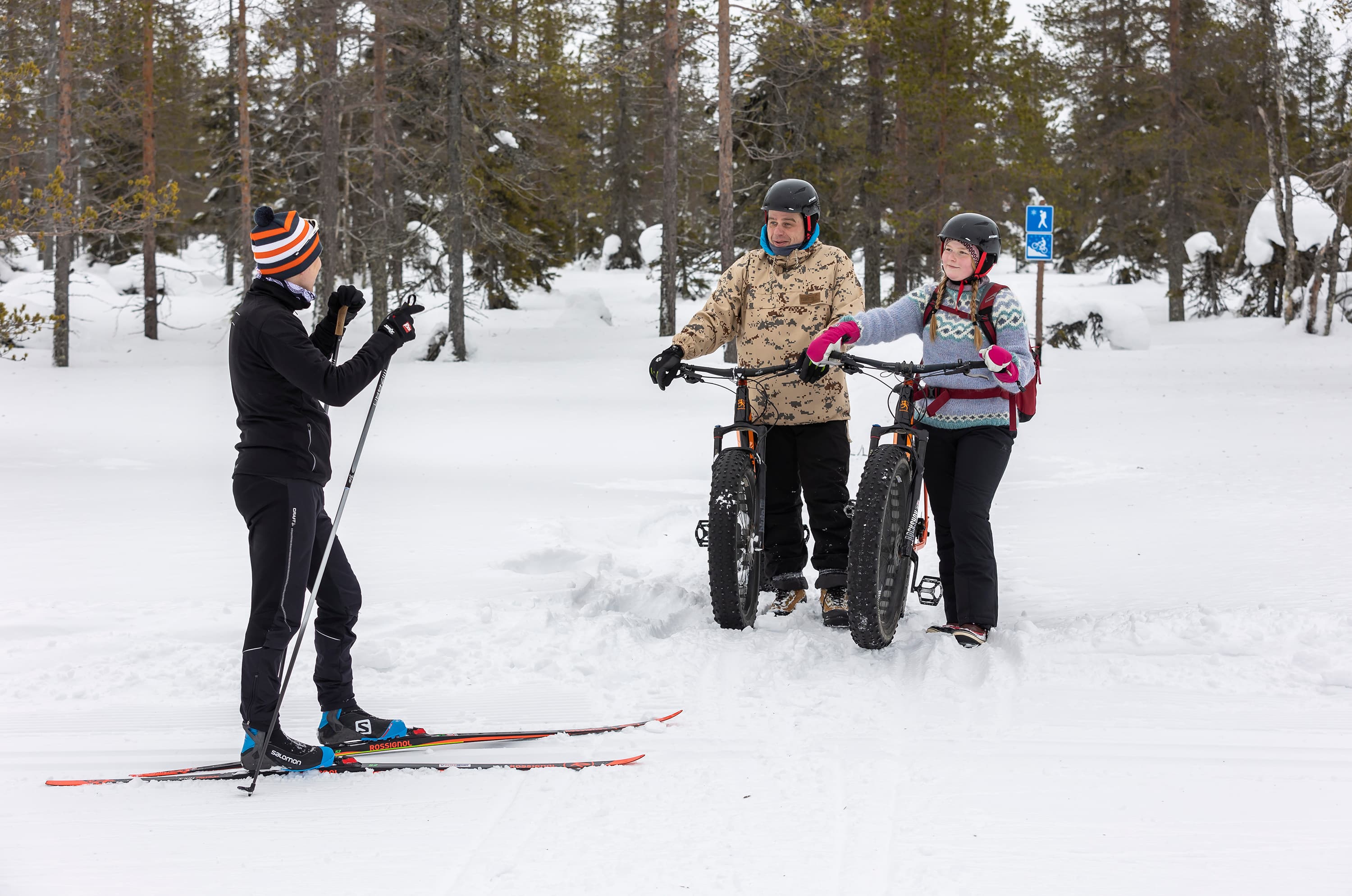 Kuva: Terhi Tuovinen Hiihto- ja pyöräilyreitti risteävät. Ladulla on seisoo hiihtäjä, pyöräilyreitillä kaksi pyöräilijää. Ihmiset juttelevat keskenään.